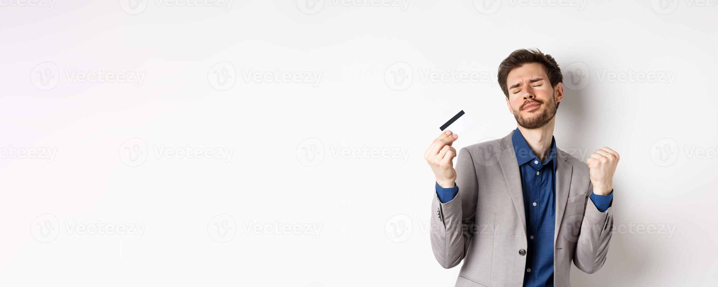 Excited successful businessman dancing with plastic credit card, raising fist pumps and smiling delighted, standing on white background in suit photo
