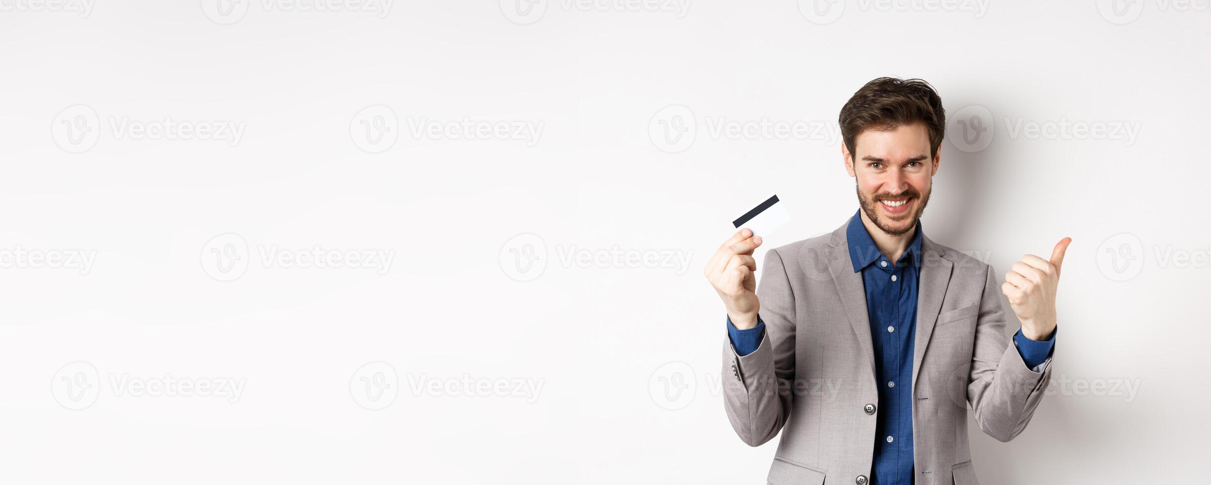 Happy bank client in business suit showing plastic credit card and thumb up, smiling satisfied, standing on white background photo