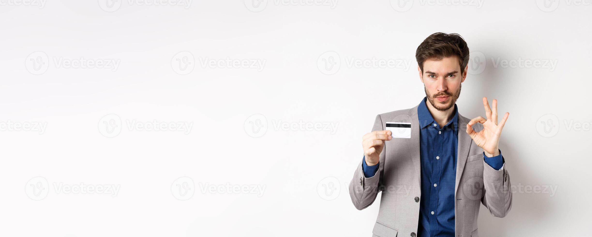 Happy male bank client in business suit showing plastic credit card and okay sign, smiling satisfied, standing on white background photo