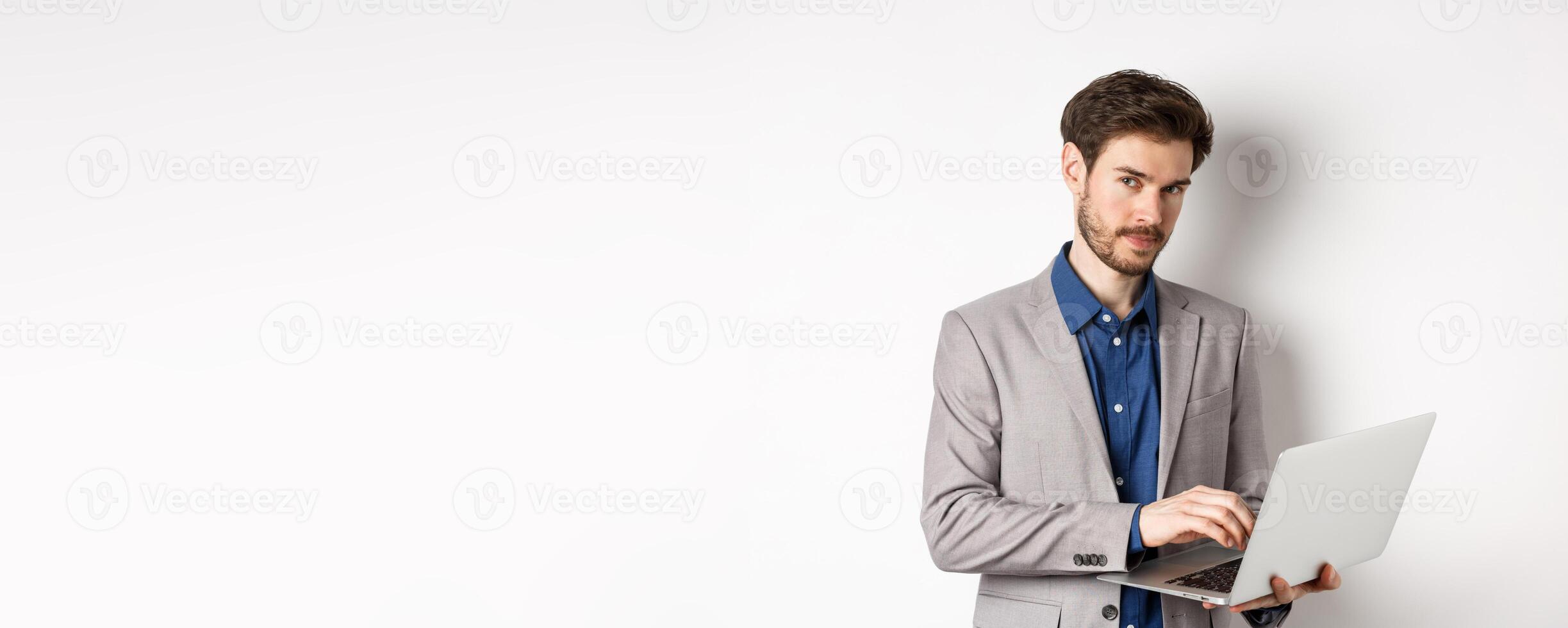 Handsome businessman in suit working on laptop, looking at camera confident, standing against white background photo