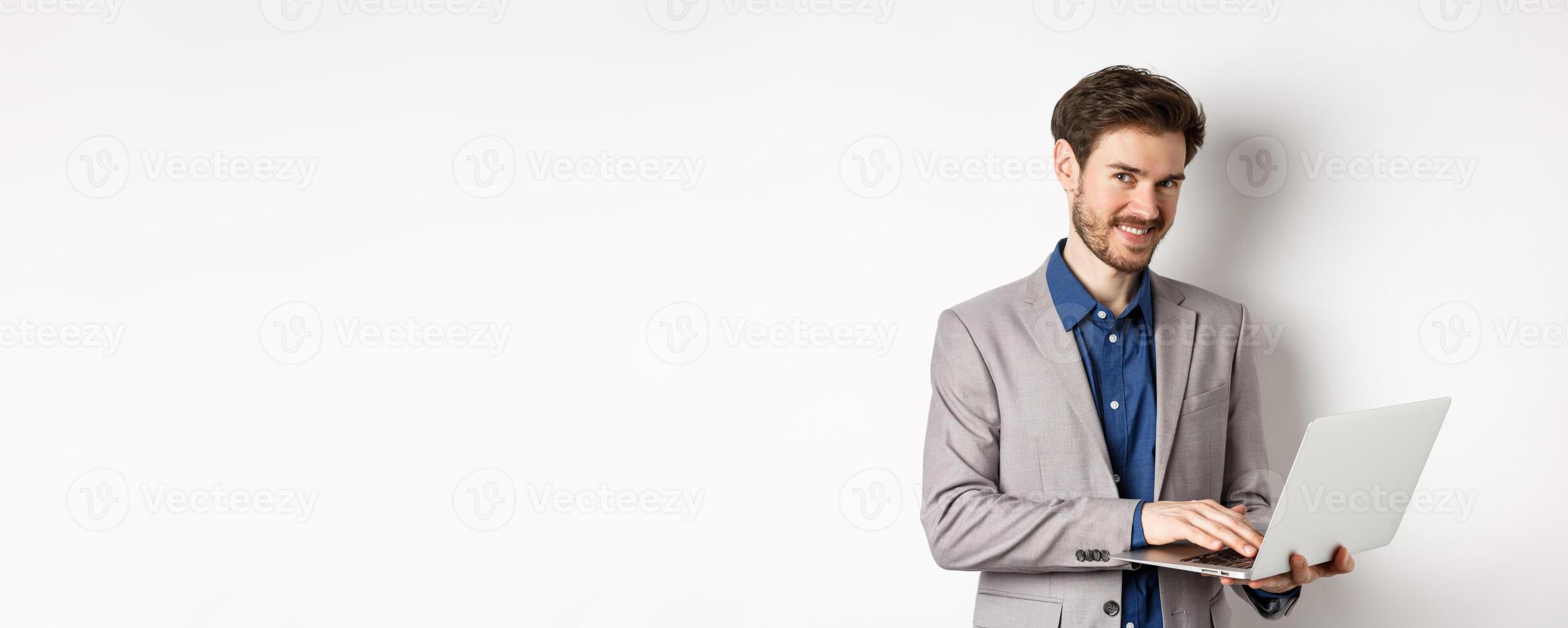Successful smiling businessman working on laptop and looking happy at camera, standing in grey suit on white background photo