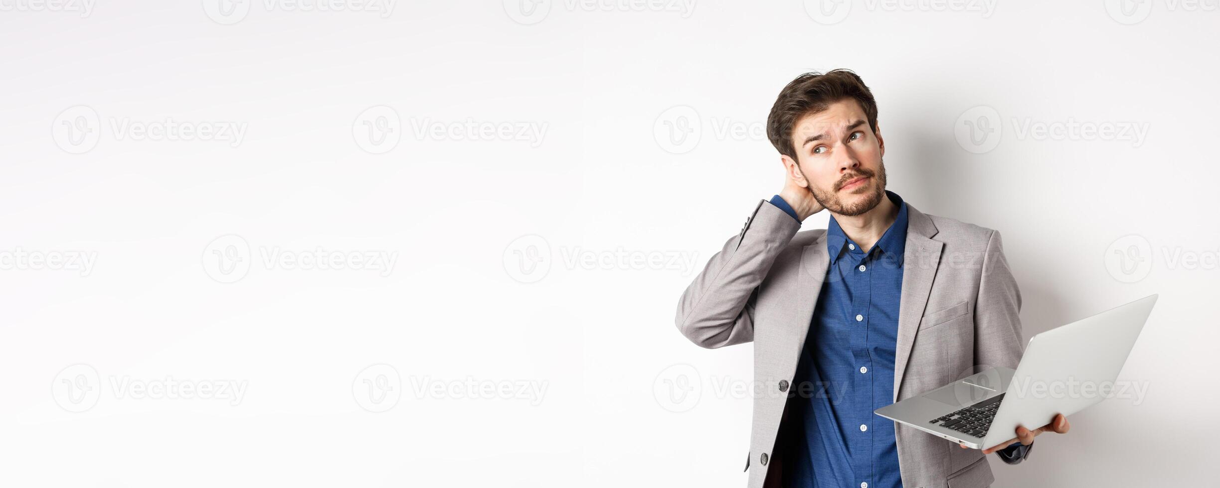 Thoughtful businessman scratch head and look away, making decision while working on laptop, standing against white background photo