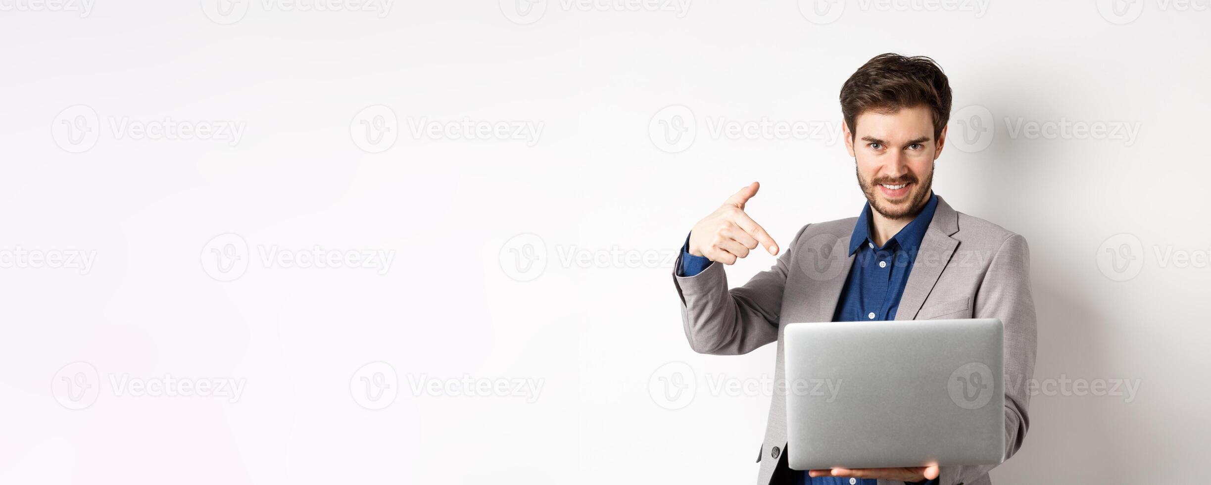 Confident young male entrepreneur working online with laptop, pointing at computer and smiling, standing on white background photo