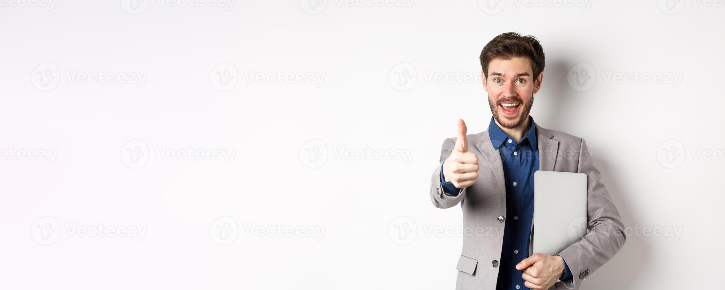 Good job. Happy smiling business man in suit holding laptop, showing thumb up to praise you, nice work gesture, standing on white background photo