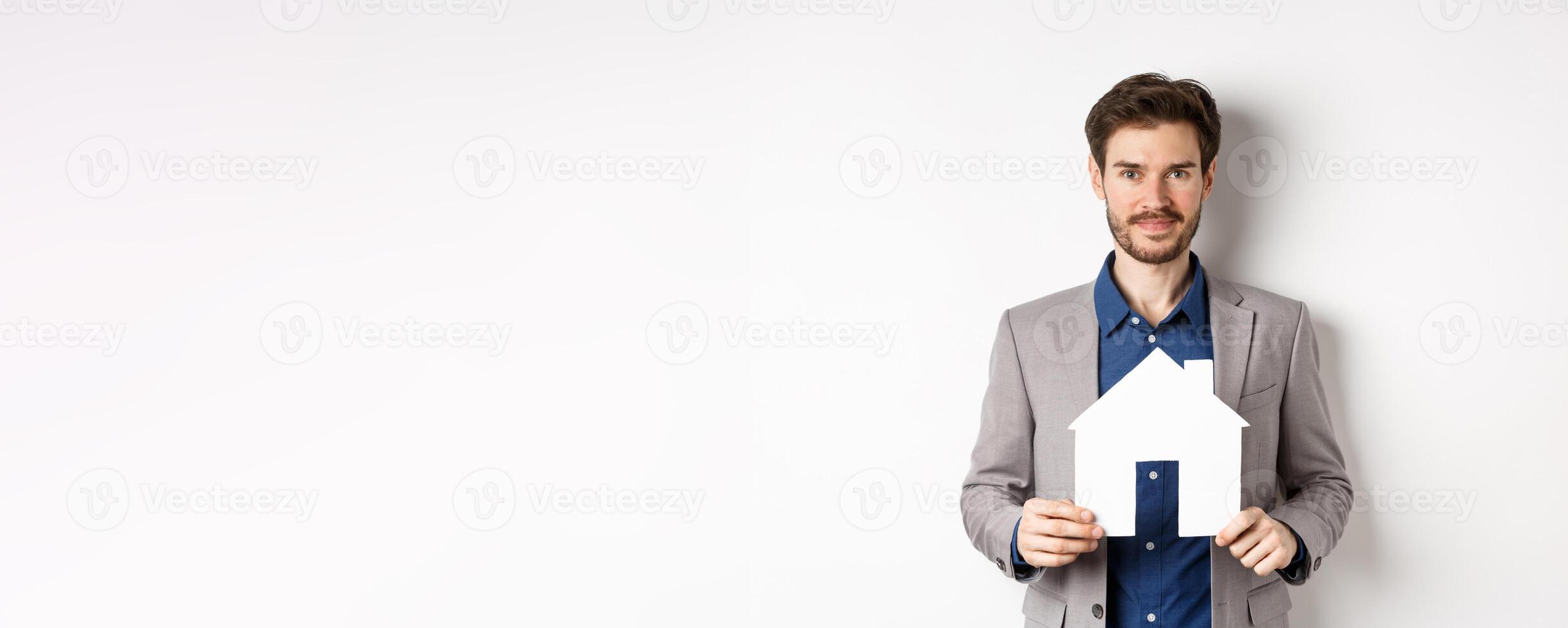 Real estate and insurance concept. Salesman in grey suit showing paper house cutout, selling property, smiling friendly at camera, white background photo