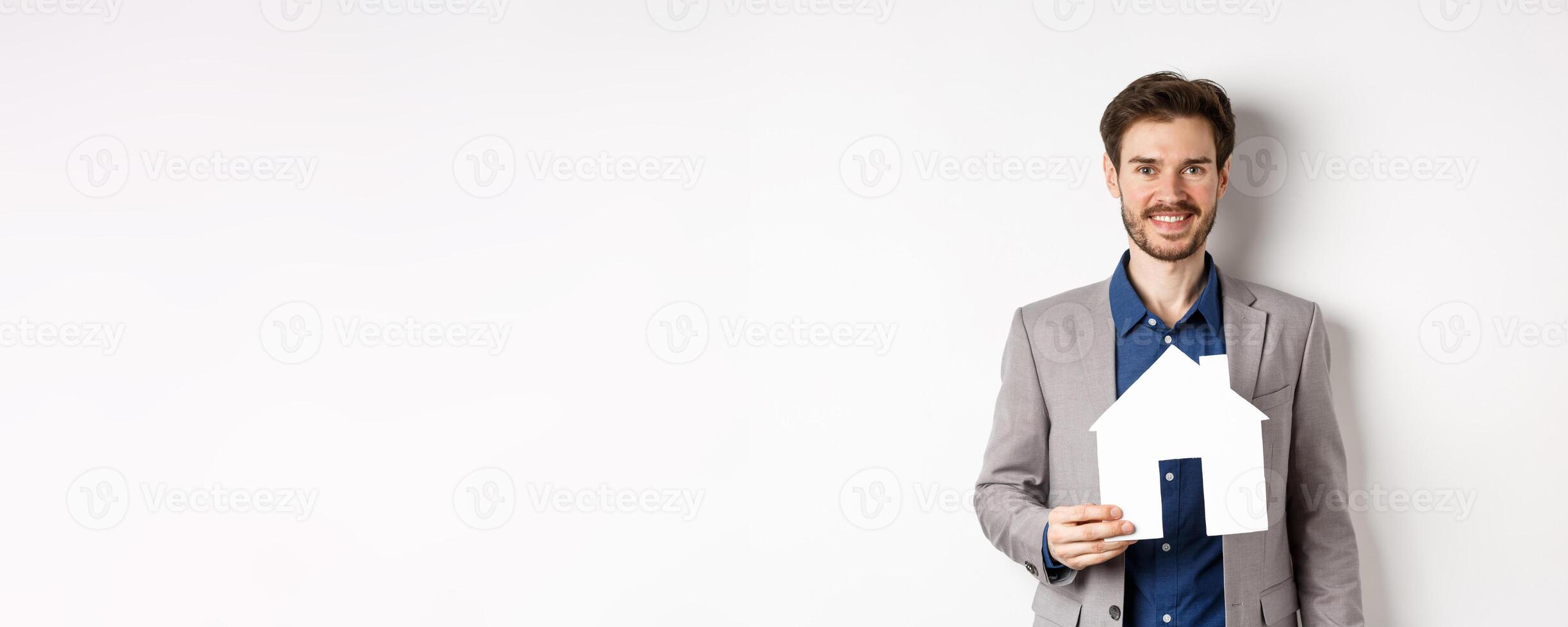 Real estate. Handsome young businessman buying property, holding paper house cutout and smiling, agency advertisement, white background photo