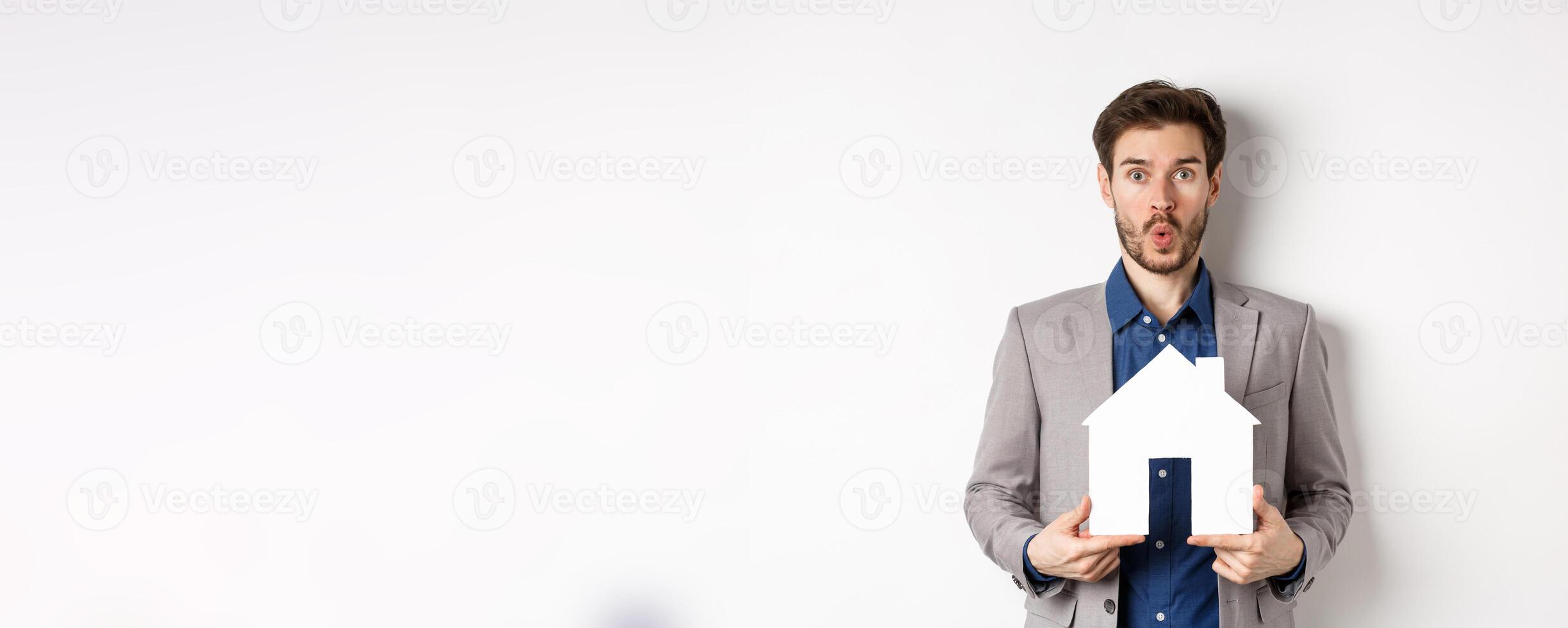 Real estate. Amazed caucasian man in suit checking out special offer for property, standing with paper house cutout and say wow at camera, white background photo