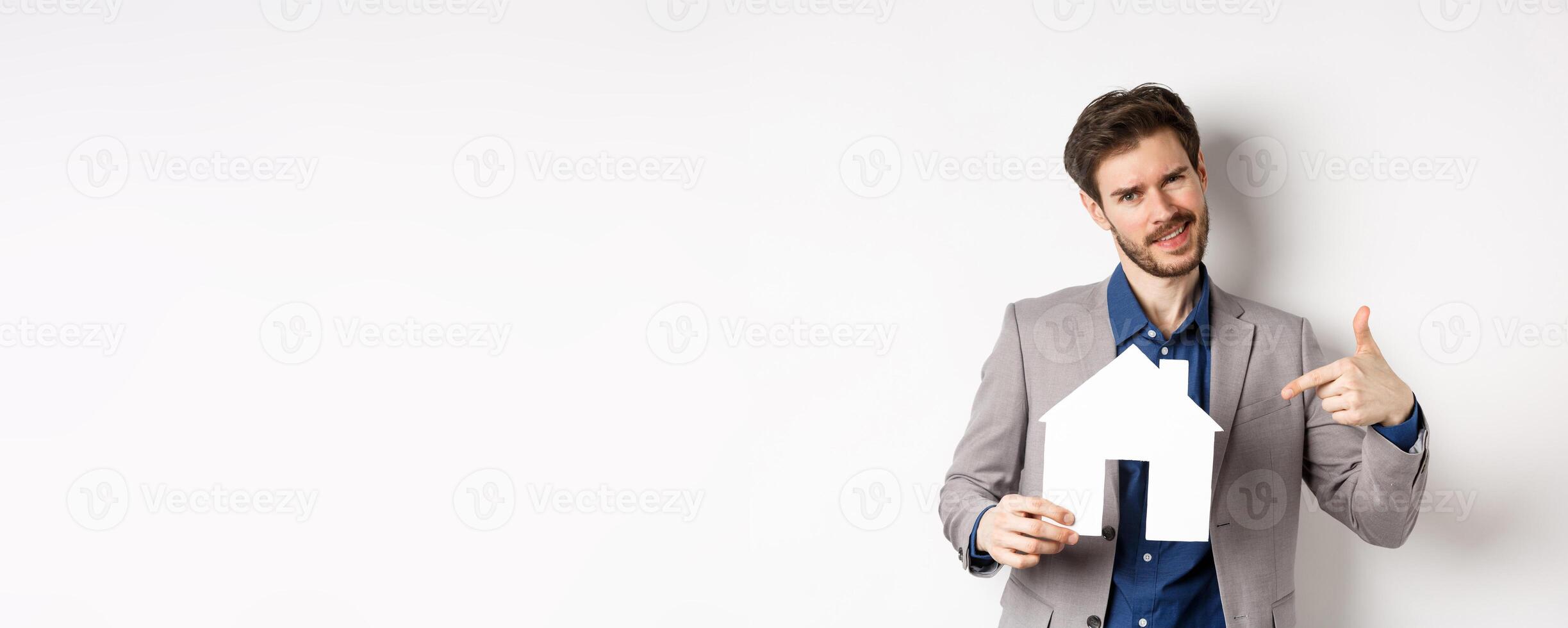Real estate and insurance concept. Salesman in grey suit showing paper house cutout, selling property, smiling at camera, white background photo