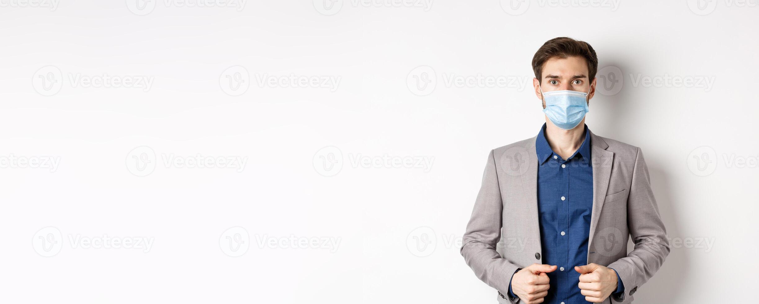 Covid-19 and office workers concept. Confident businessman in medical mask and suit looking at camera, standing on white background photo