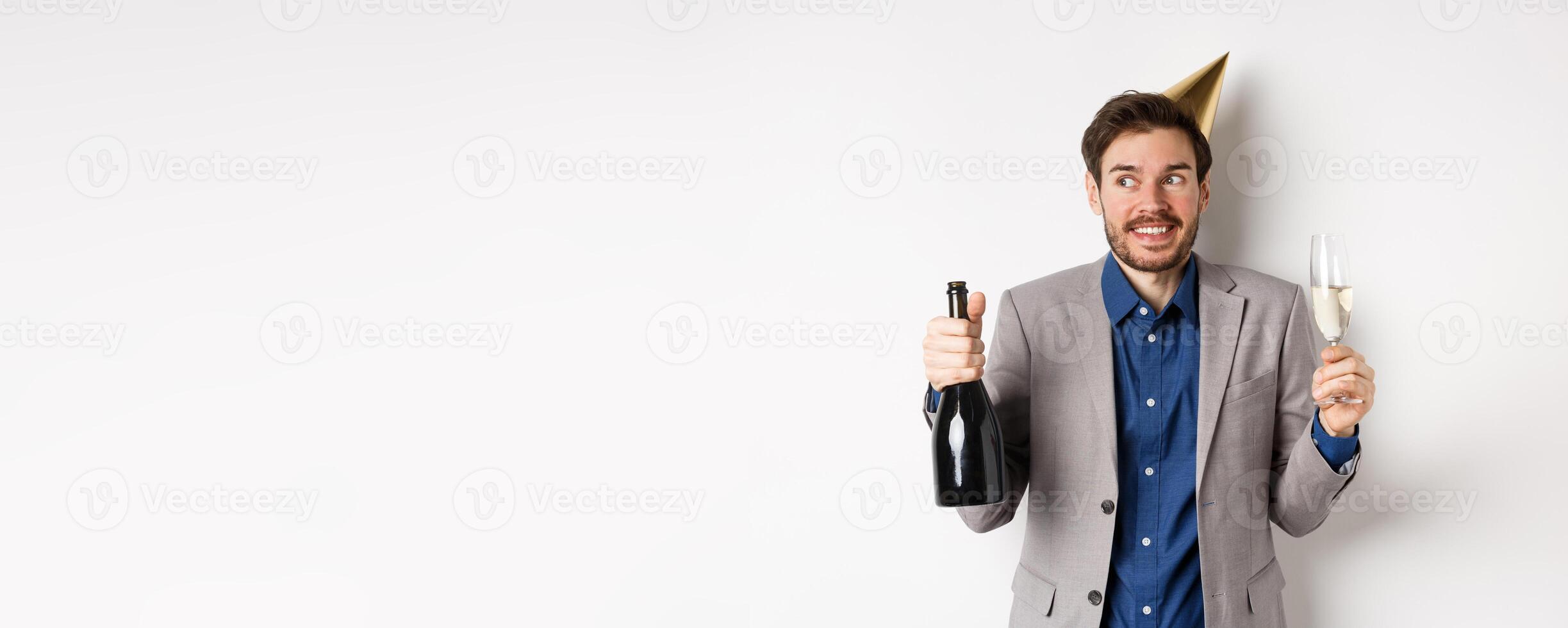 Celebration and holidays concept. Happy birthday guy in suit and party hat drinking champagne, holding bottle and glass, smiling and looking aside photo