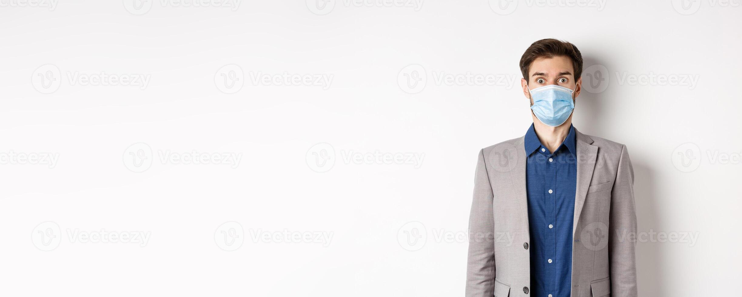 Health, pandemic and business concept. Surprised guy in suit and medical mask raising eyebrows, stare shocked at camera, white background photo