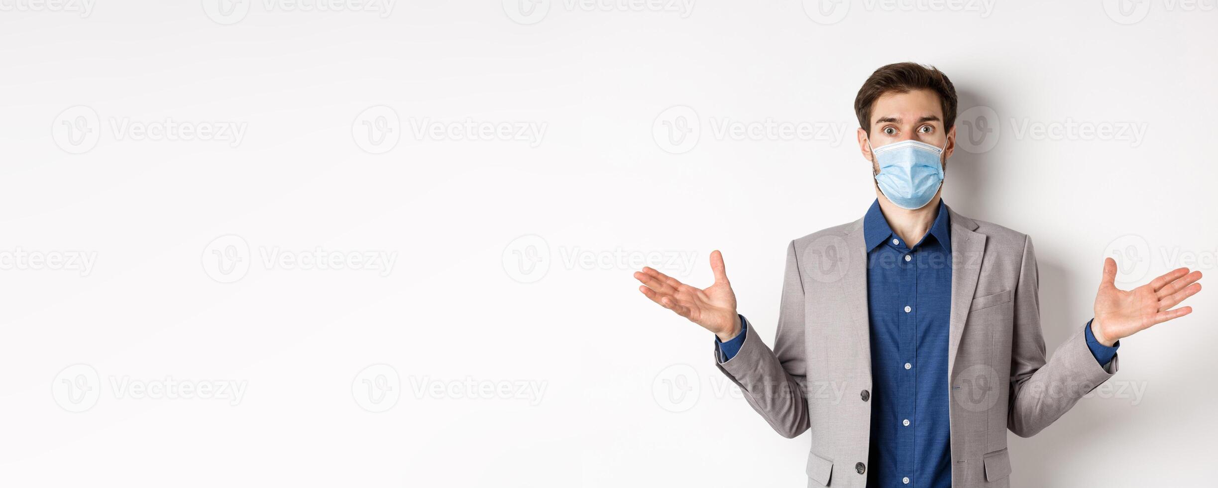 Health, pandemic and business concept. Confused businessman in medical mask and suit spread hands sideways and look puzzled, cant understand, white background photo