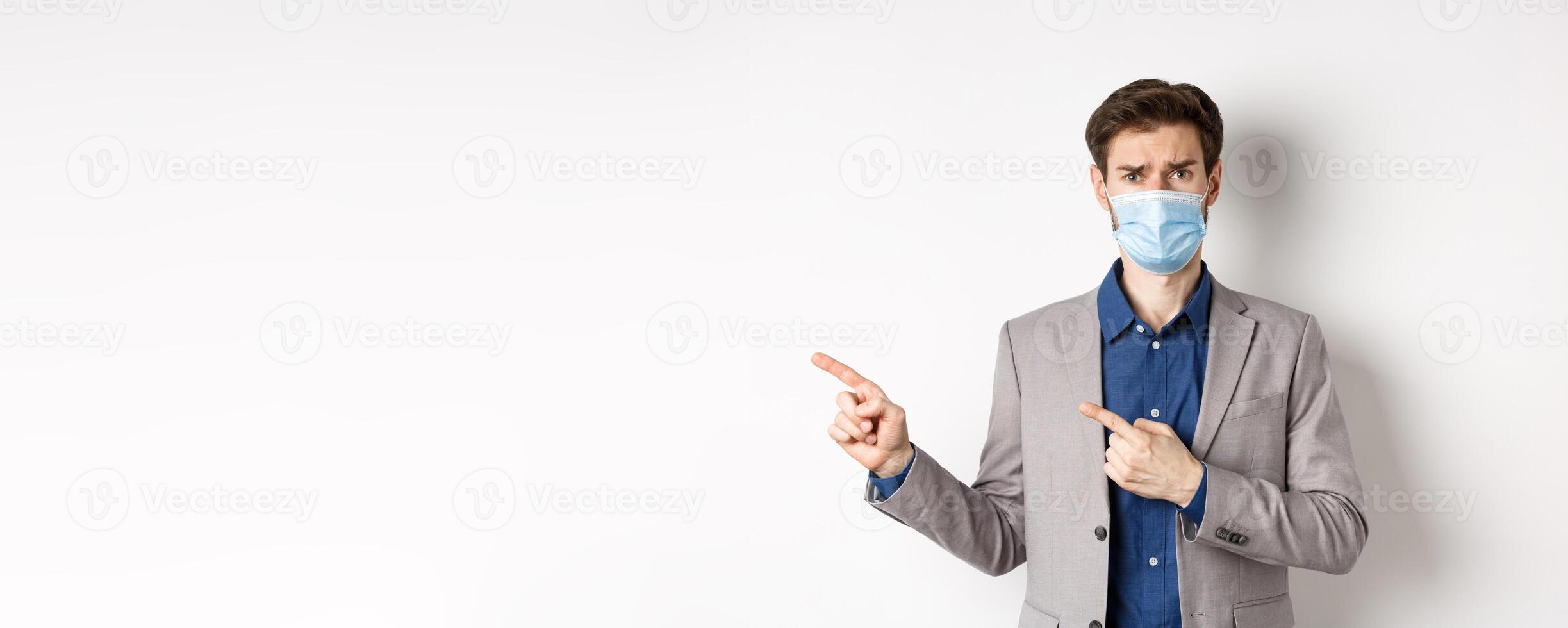 Covid-19, pandemic and business concept. Upset man in medical mask and office suit, frowning sad and pointing left at banner, standing on white background photo