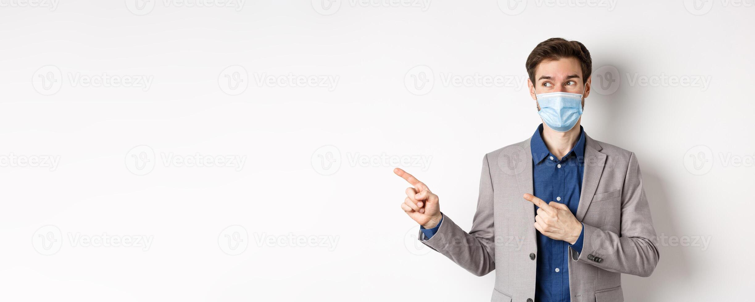 Covid-19, pandemic and business concept. Excited young man in medical mask and suit looking left, pointing at logo with pleased face, white background photo