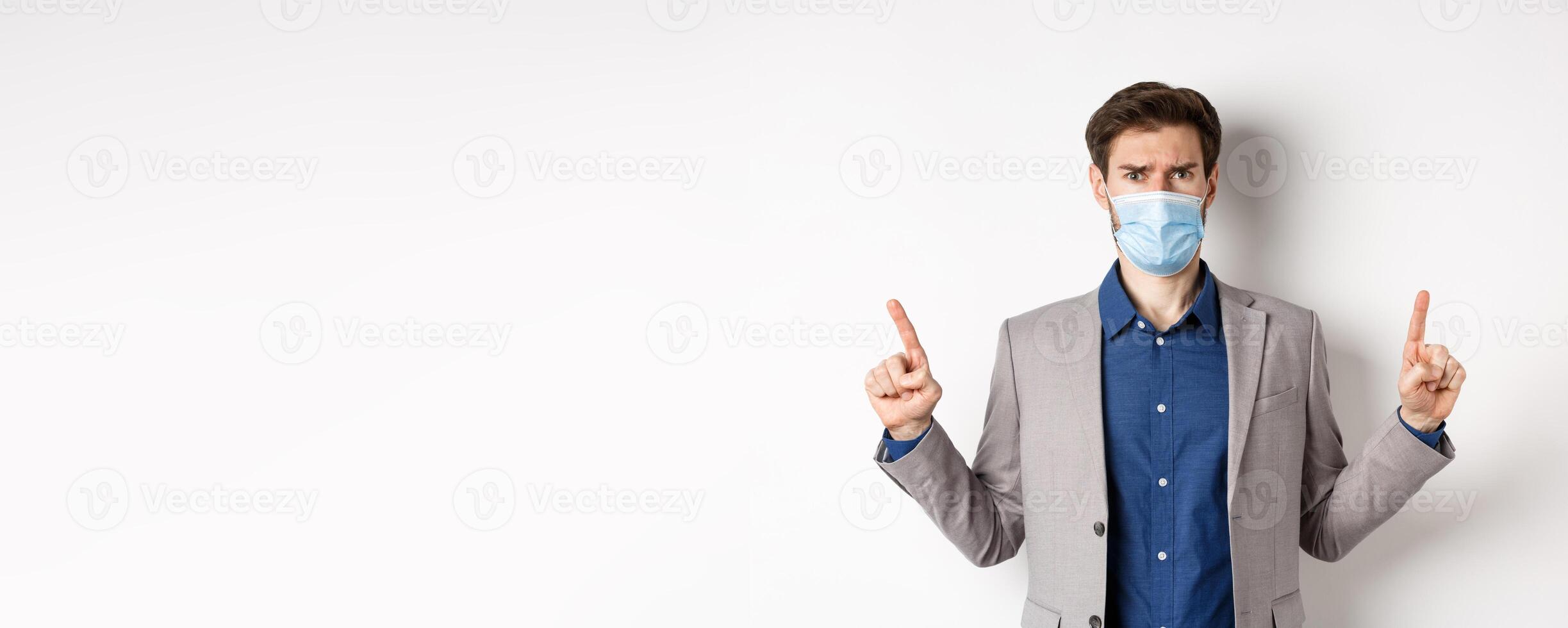 Covid-19, pandemic and business concept. Disappointed frowning guy in suit and medical mask pointing fingers up, complaining on unfair situation, white background photo
