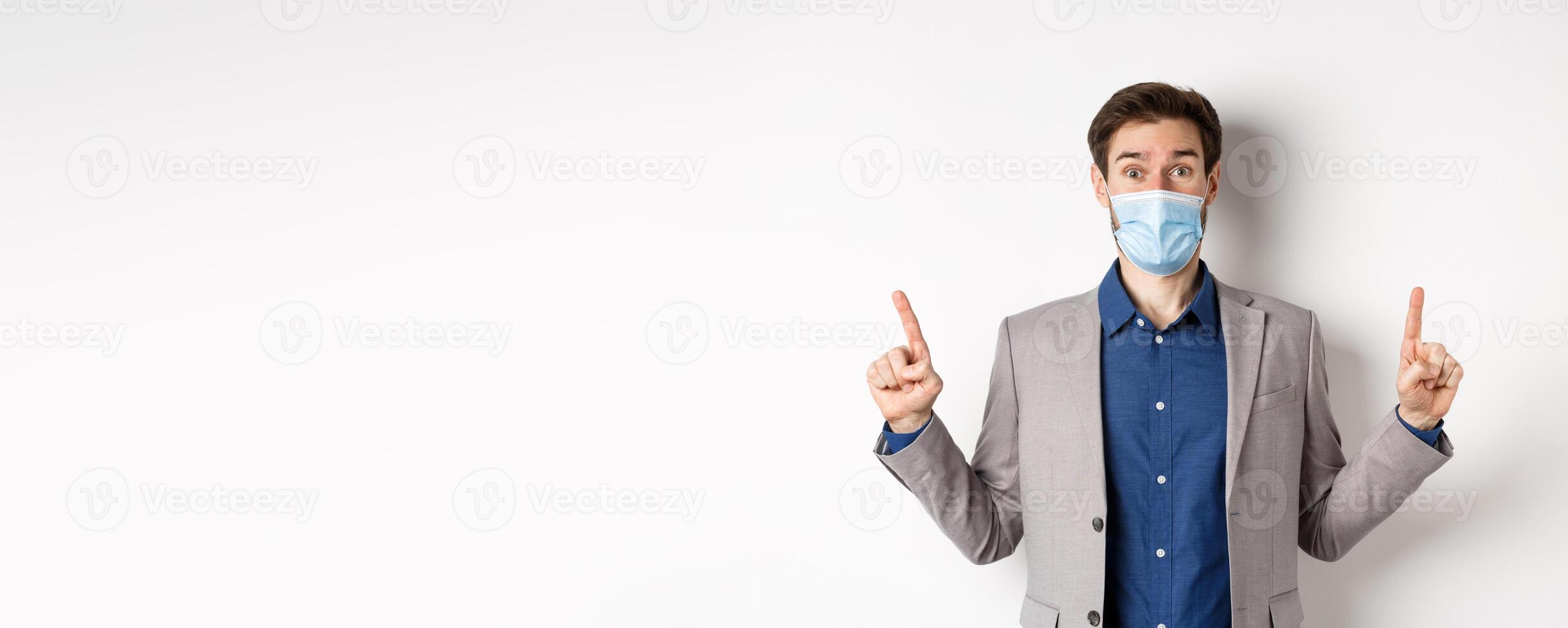 Covid-19, pandemic and business concept. Excited businessman in sterile medical mask and suit pointing fingers up, white background photo