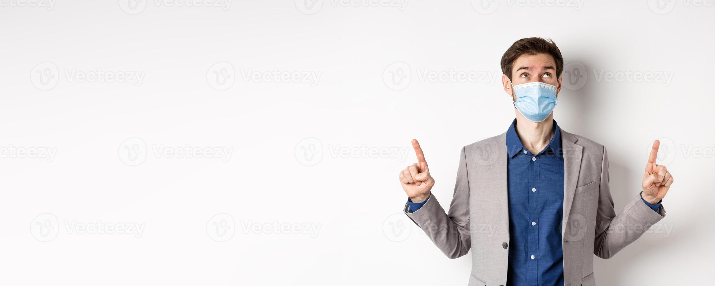 Covid-19, pandemic and business concept. Handsome businessman in medical mask and suit, looking and pointing up with dreamy face, white background photo