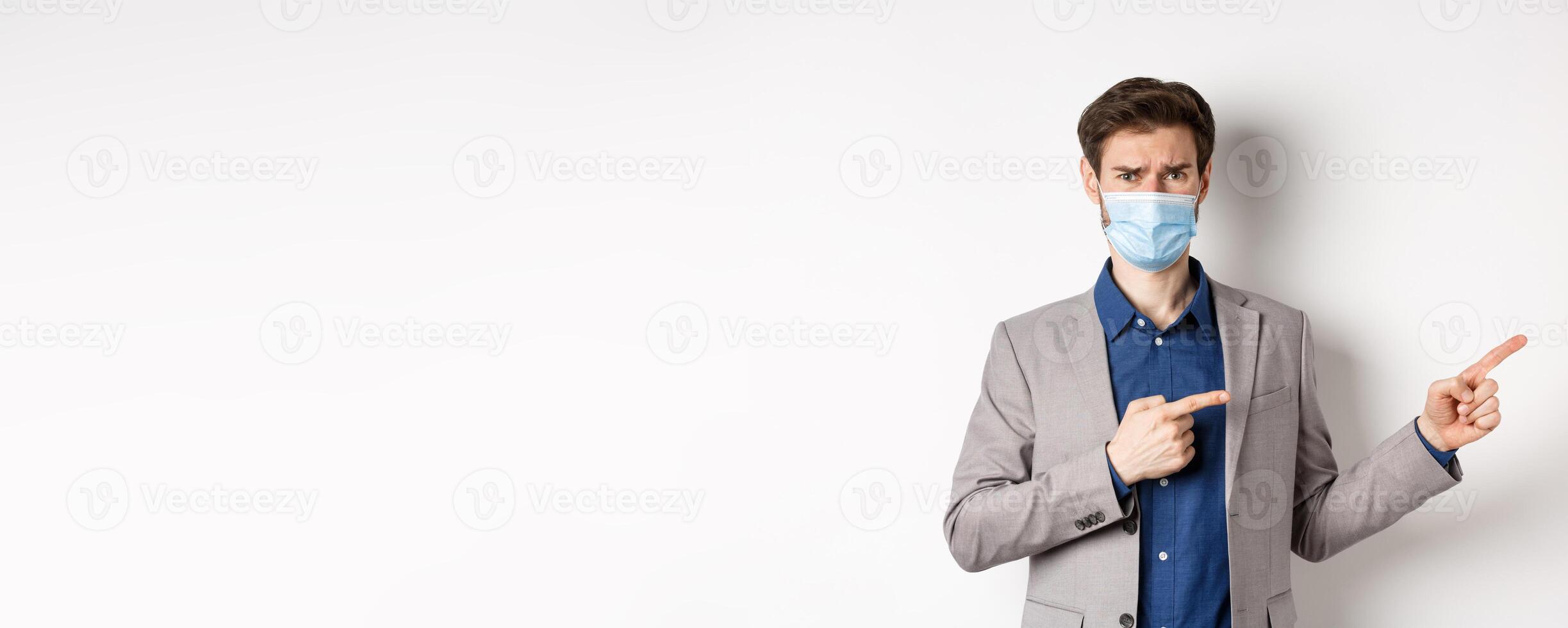 Covid-19, pandemic and business concept. Disappointed and reluctant man in medical mask frowning, pointing fingers right at something disgusting or ugly, white background photo
