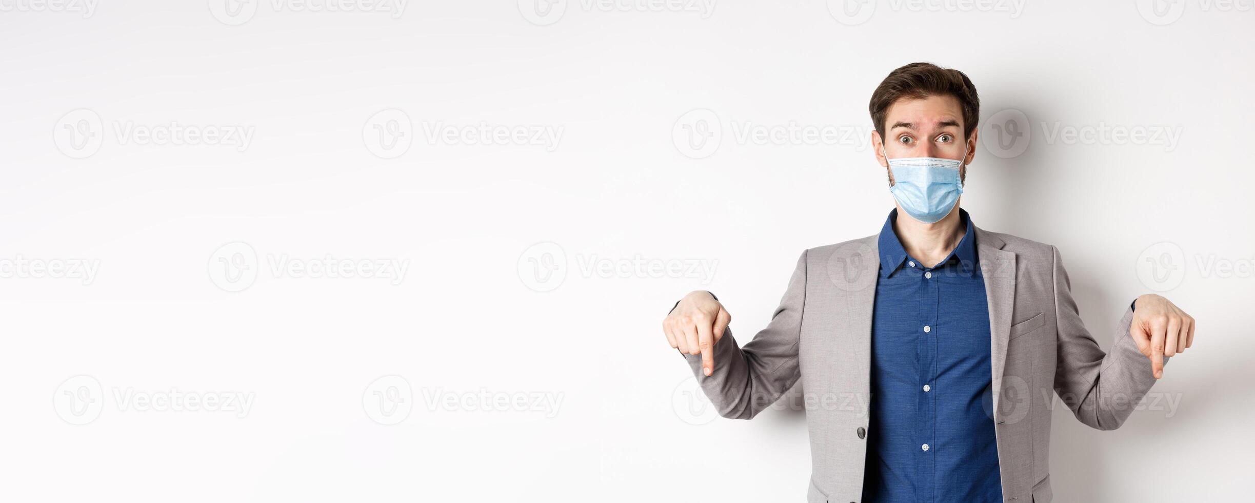 Covid-19, pandemic and business concept. Surprised male ceo manager in medical mask and suit pointing fingers down, looking amused, white background photo
