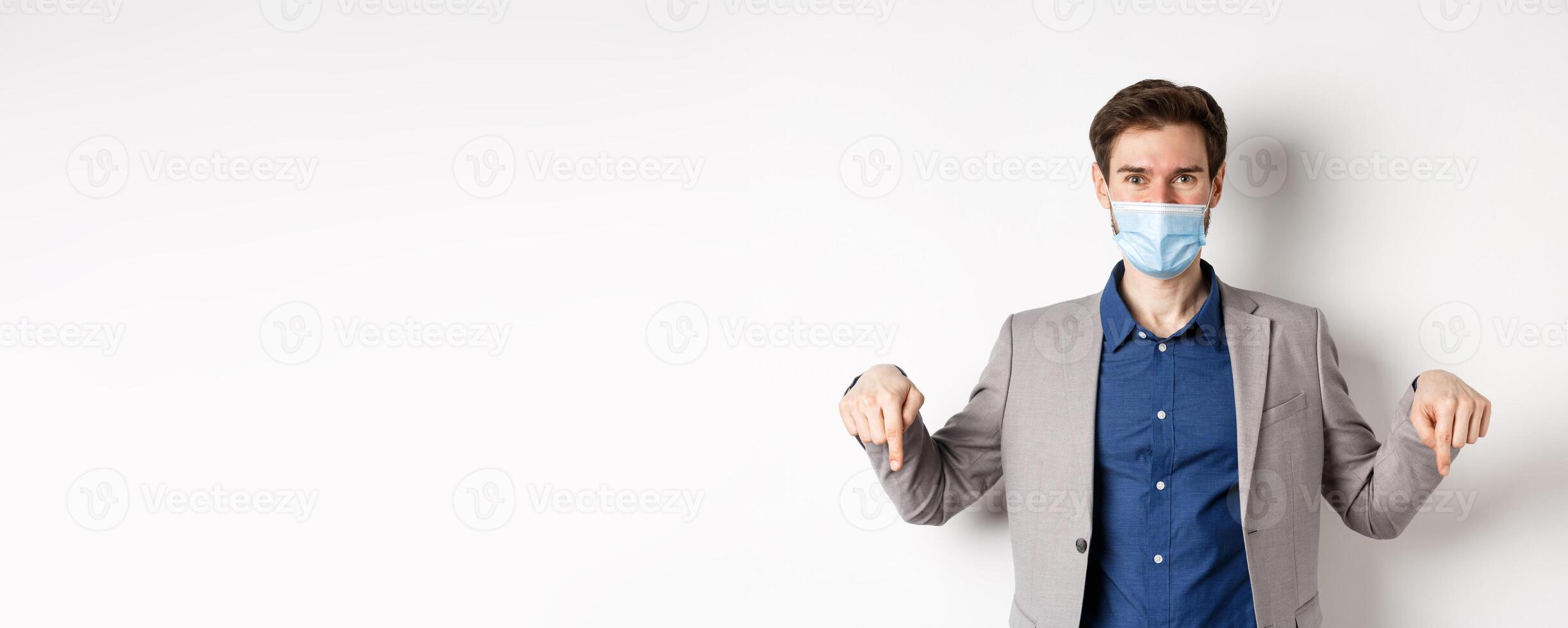 Covid-19, pandemic and business concept. Handsome businessman in medical mask and suit, pointing fingers down and showing company logo, white background photo