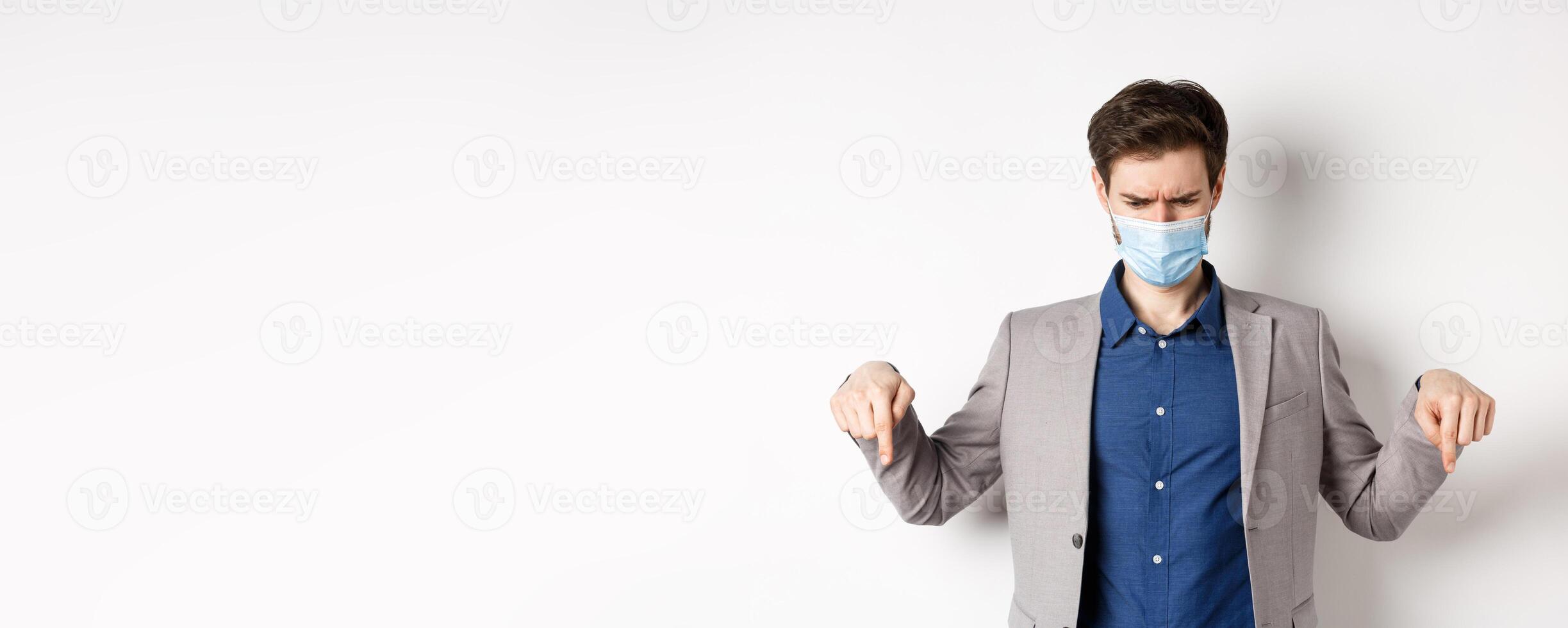 Covid-19, pandemic and business concept. Confused and displeased businessman in medical mask frowning, pointing fingers down and looking disappointed, white background photo