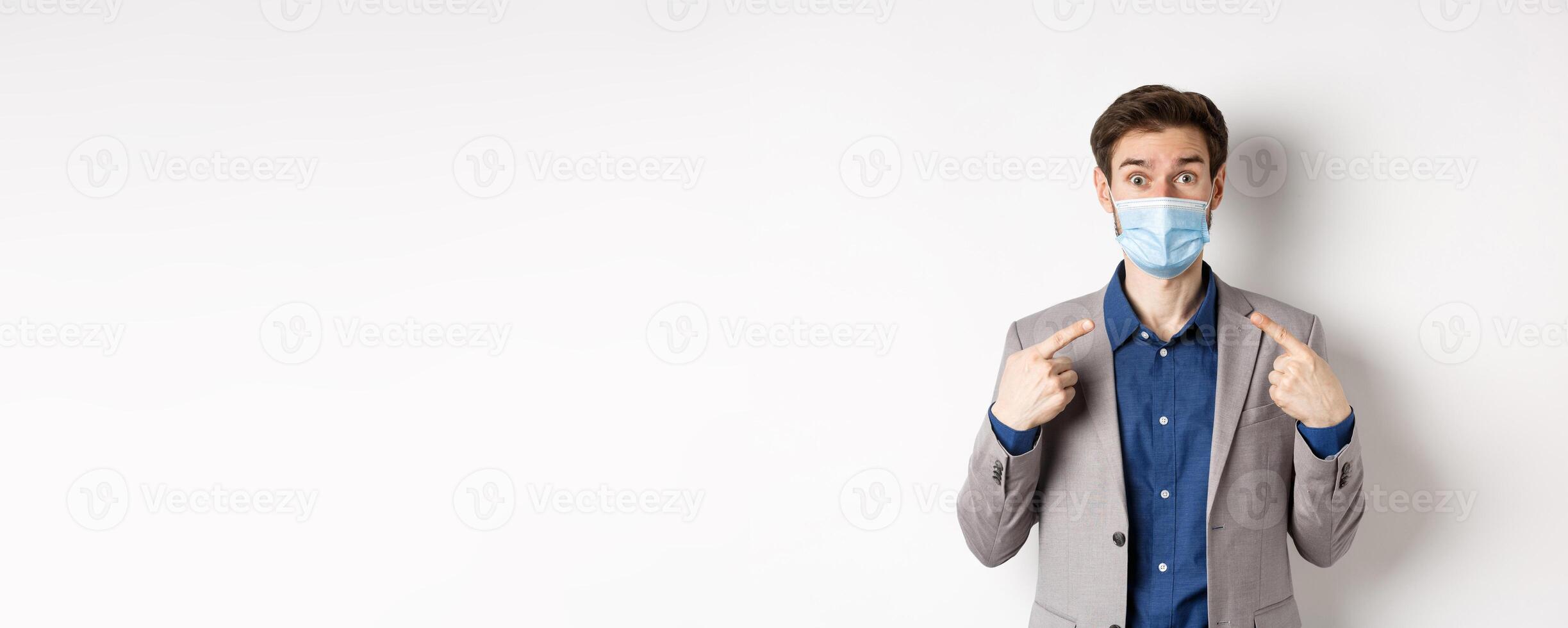 Covid-19, pandemic and business concept. Excited businessman in suit pointing at his medical mask and looking at camera, white background photo