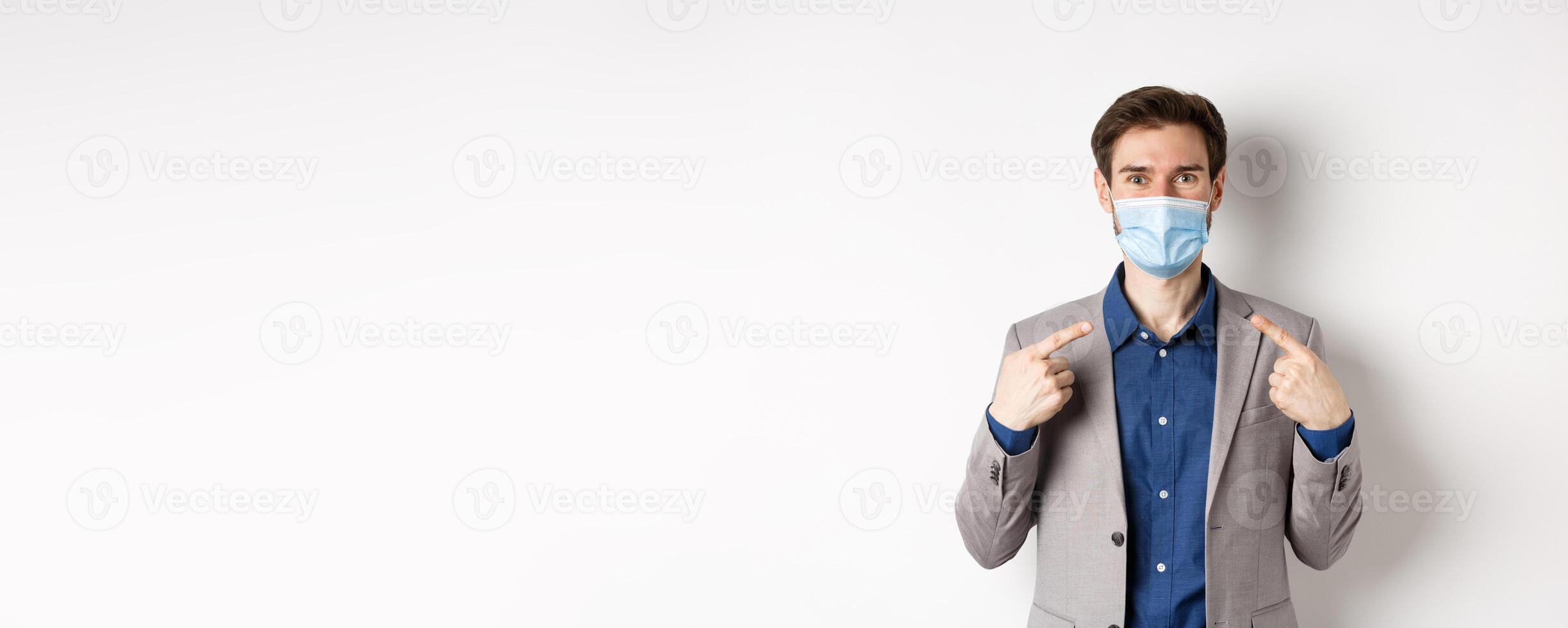 Covid-19, pandemic and business concept. Excited businessman in suit pointing at his medical mask and smiling with eyes, white background photo