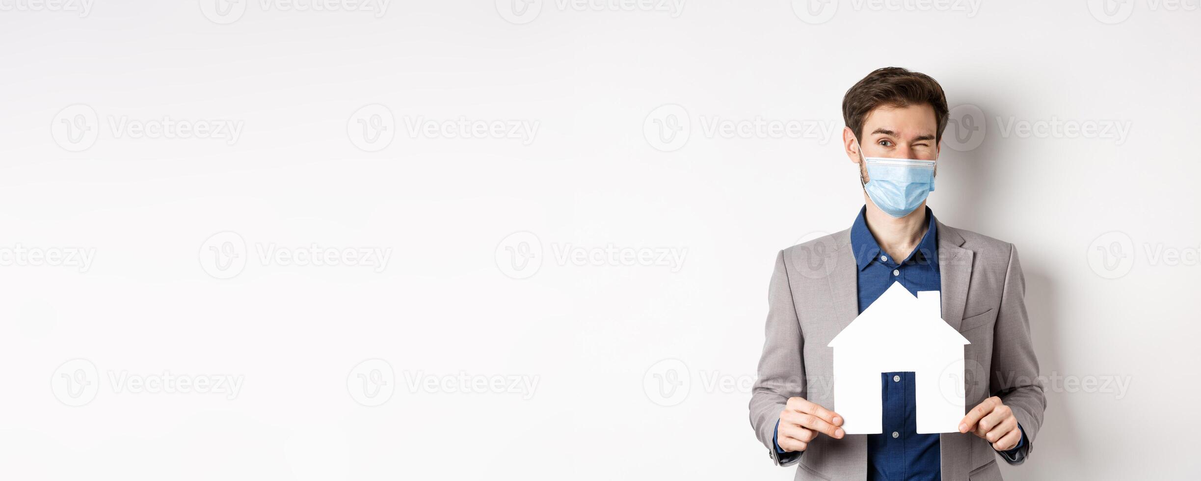 Real estate and covid-19 concept. Cheerful young man in medical mask and suit winking at camera, advertising new flats, showing paper house cutout photo