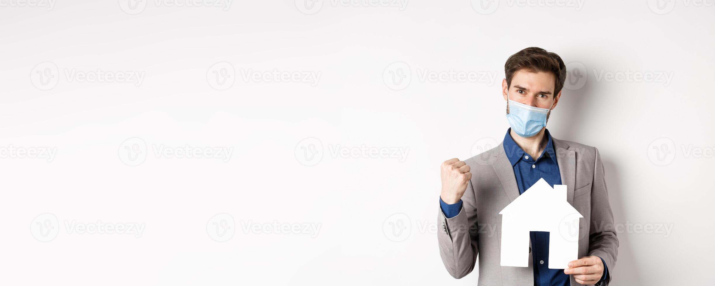 Real estate and covid-19 concept. Excited man in medical mask and suit motivated to buy house, holding paper home cutout and say yes, white background photo