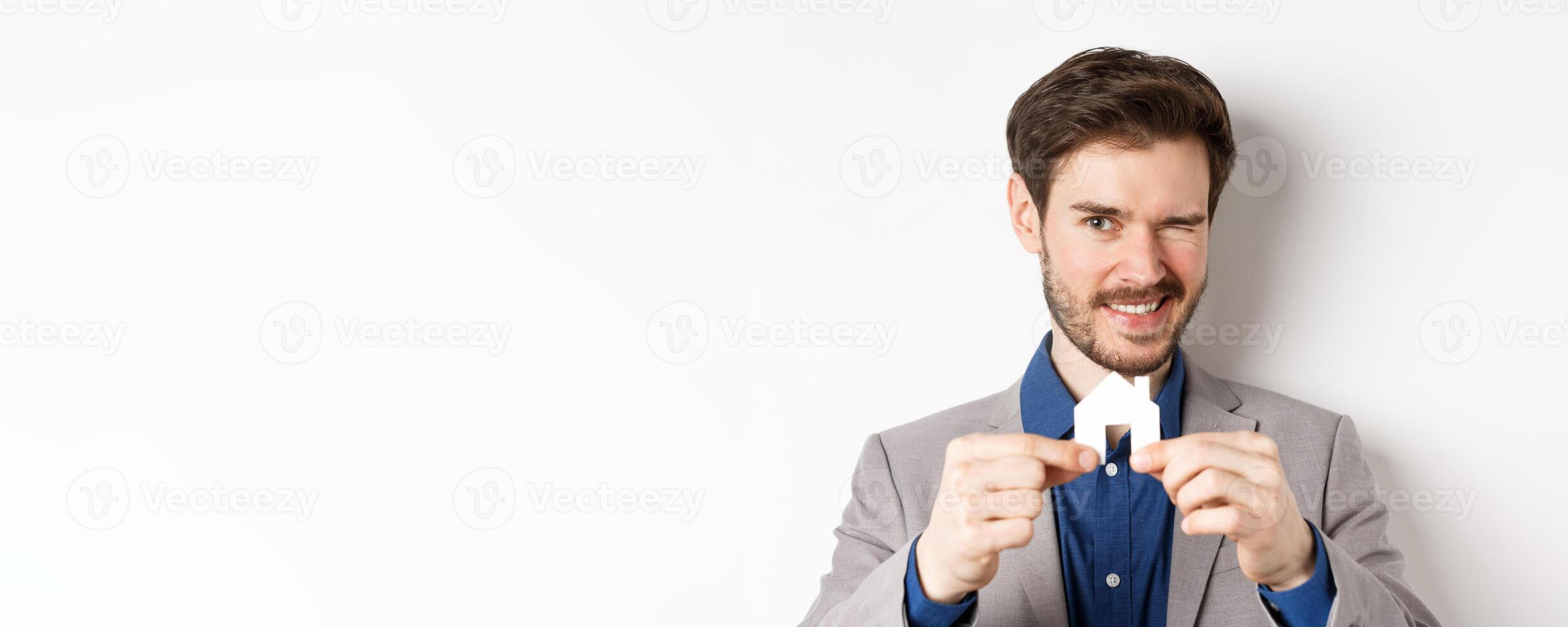 Real estate and insurance concept. Handsome man in suit winking and smiling, showing small paper house cutout, standing on white background photo