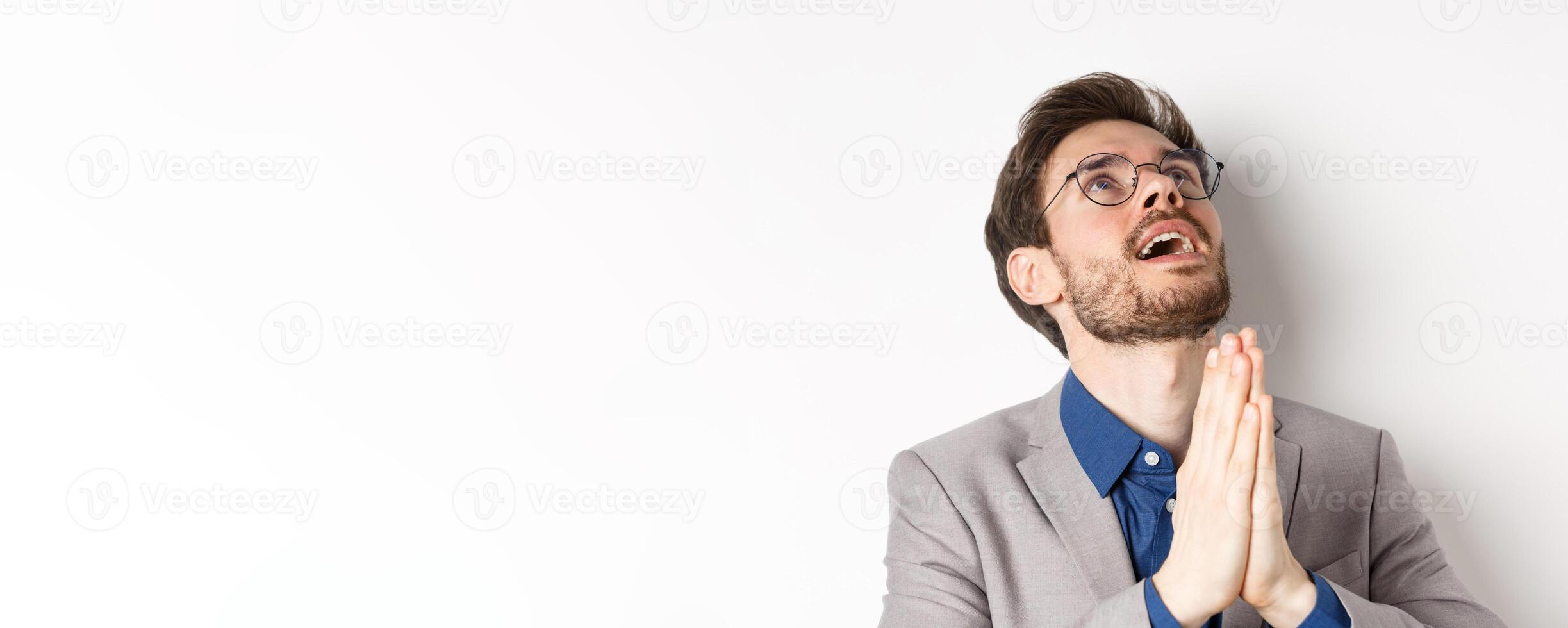 Close-up of handsome bearded man in glasses and suit looking up and praying god, begging lord for something, standing on white background photo