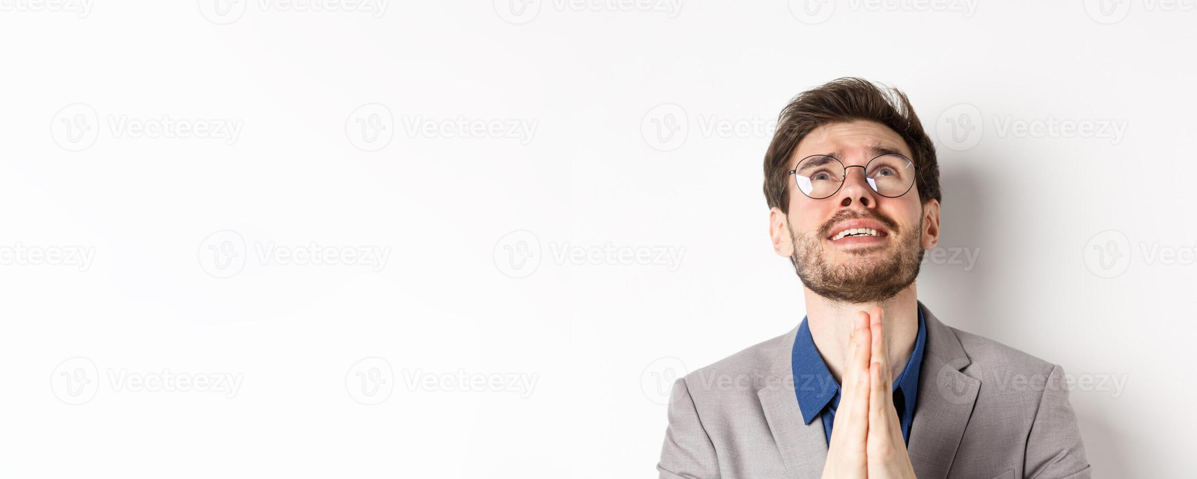 Nervous hopeful man in glasses and suit begging god, asking please and shaking hands in pray, white background photo