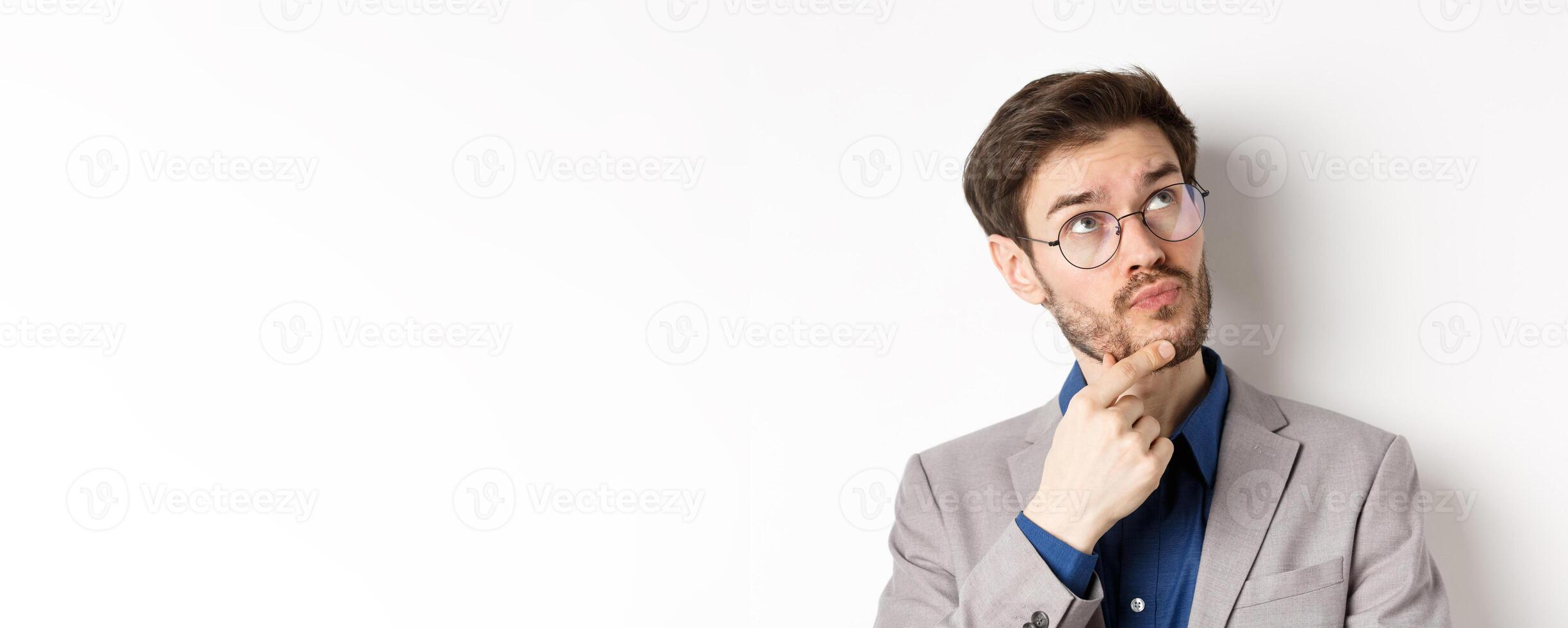 Thoughtful businessman with beard and glasses making decision, looking up and thinking, standing in suit with pensive face, white background photo