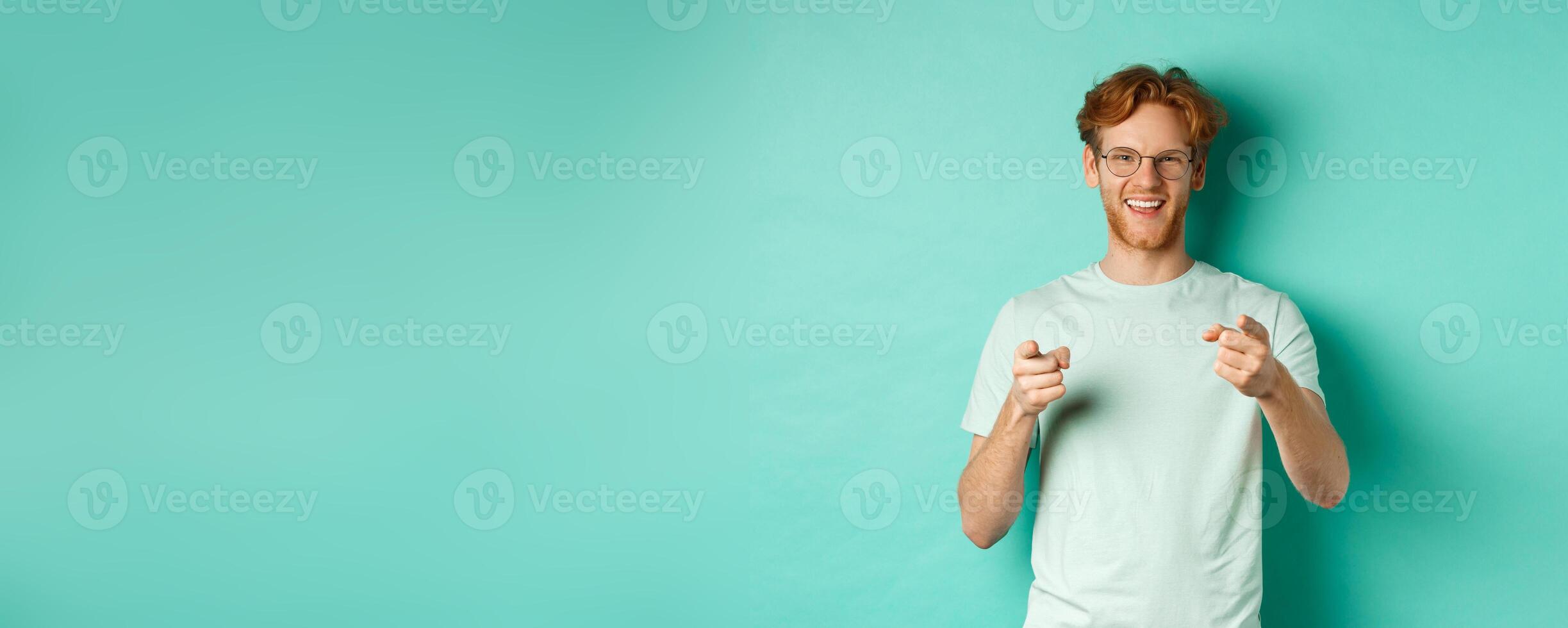 Handsome young man with ginger hair, wearing glasses and t-shirt, pointing finger at camera and smiling, choosing you, congratulating or praising, standing over mint background photo
