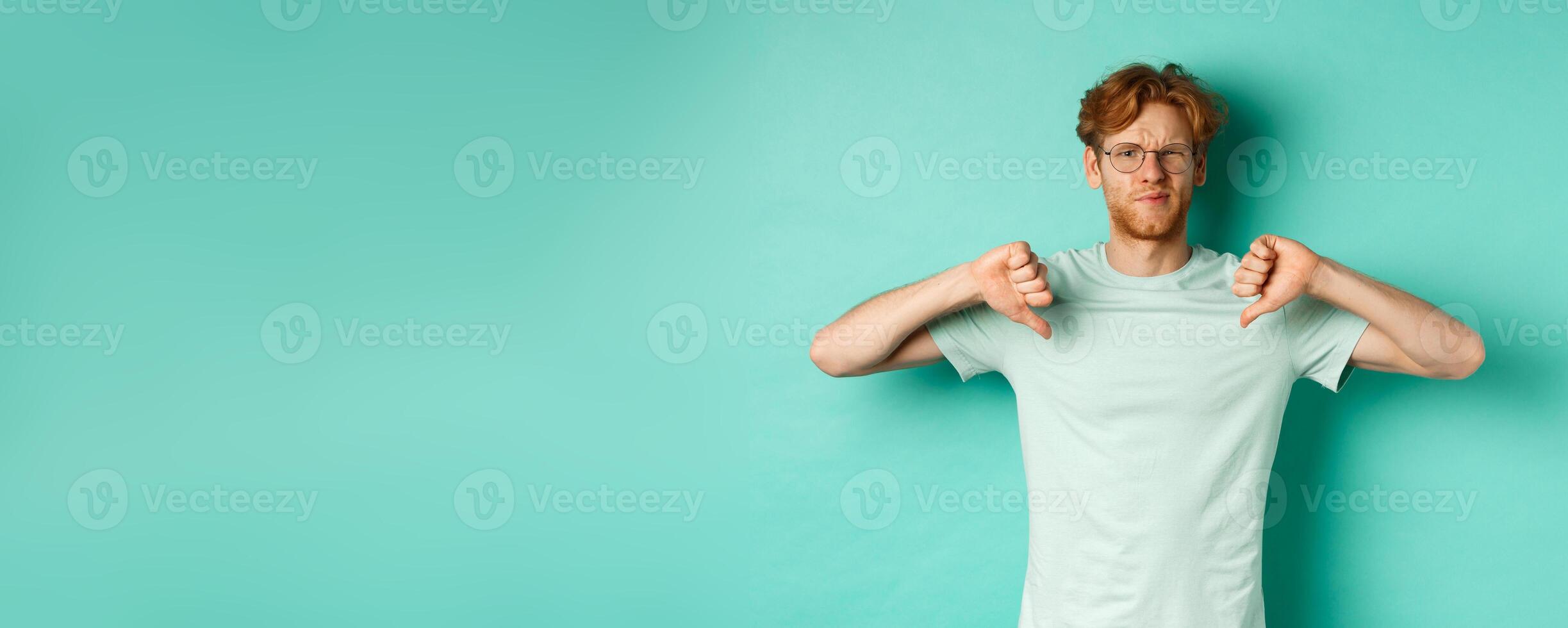 Disappointed caucasian guy with red messy hair and glasses showing thumbs down, dislike something, frowning upset and displeased, standing over mint background photo