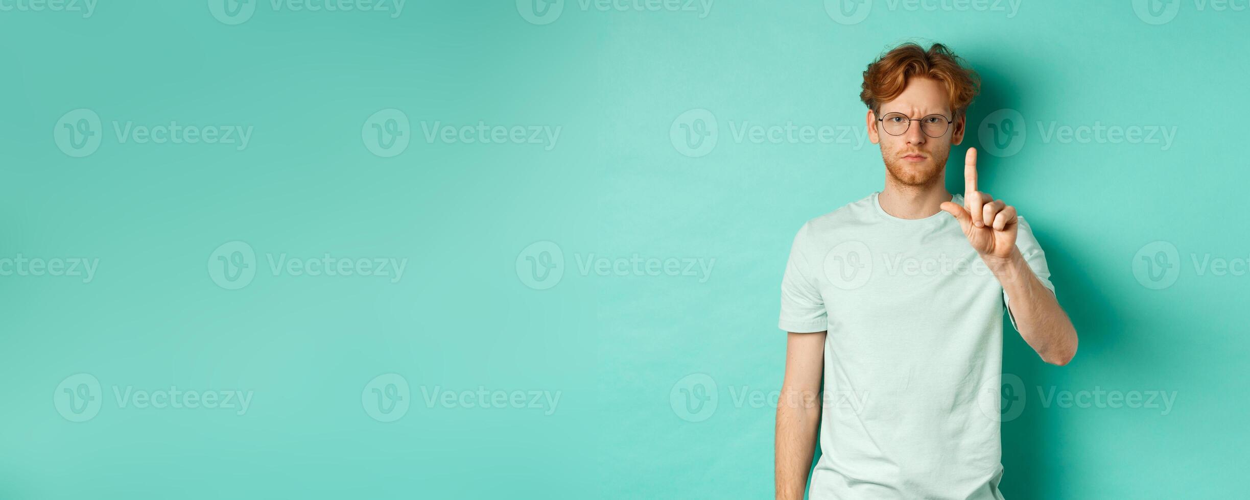 Angry and serious young man with red hair, wearing glasses, showing stop gesture, telling no, shaking finger with disapproval, standing over mint background photo