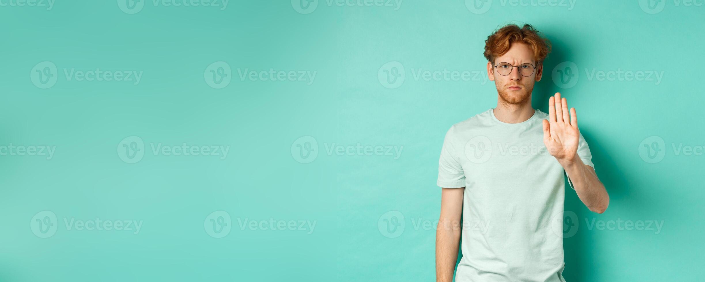 Angry and serious young man with red hair, wearing glasses, showing stop gesture, telling no, disapprove and prohibit something bad, standing over turquoise background photo