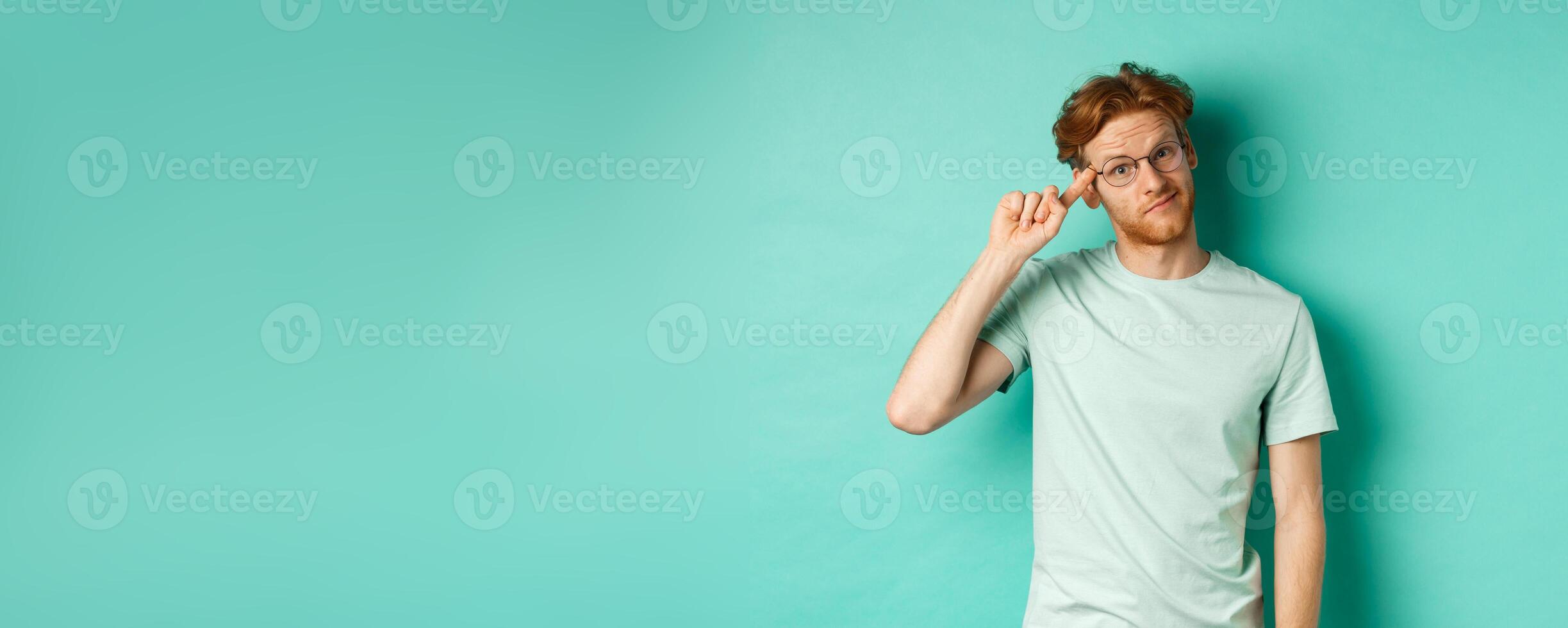 Skeptical redhead man scolding someone stupid or crazy, pointing finger at head and staring at camera, standing over mint background photo