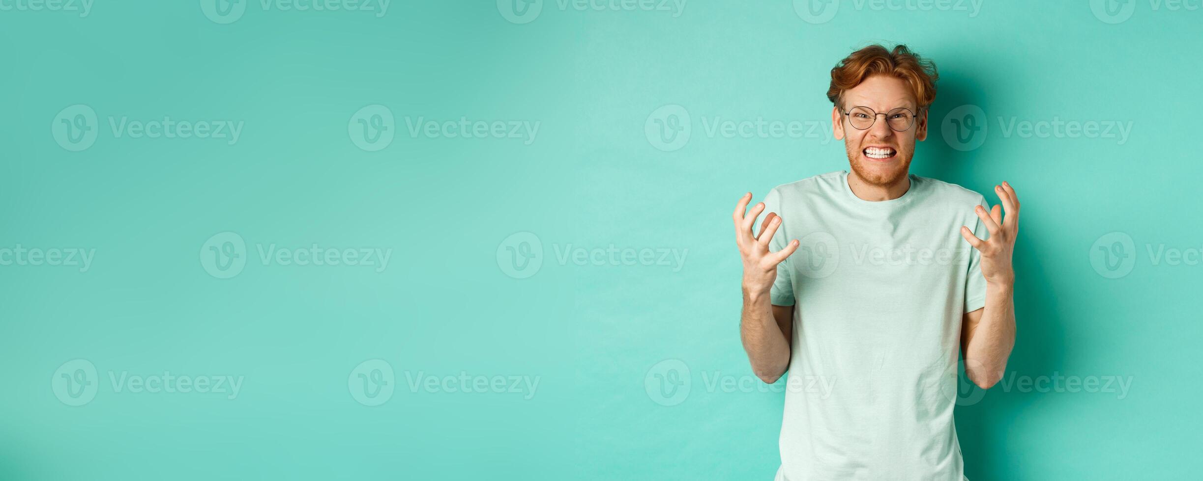 Portrait of distressed and angry redhead guy losing temper, shouting and shaking hands outraged, staring with furious face at camera, standing over mint background photo