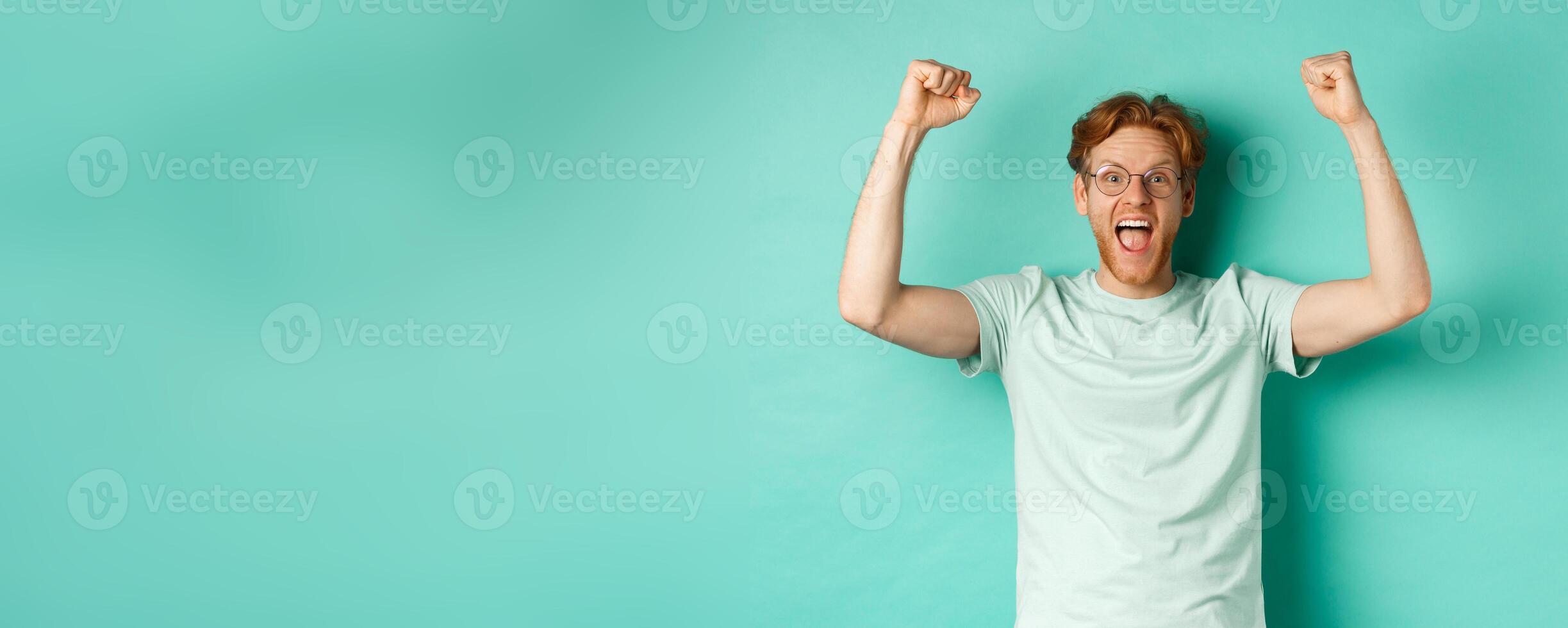 Young redhead man feeling like champion, raising hands up in fist pump gesture and shouting yes with joy, winning prize, triumphing of success, standing over mint background photo