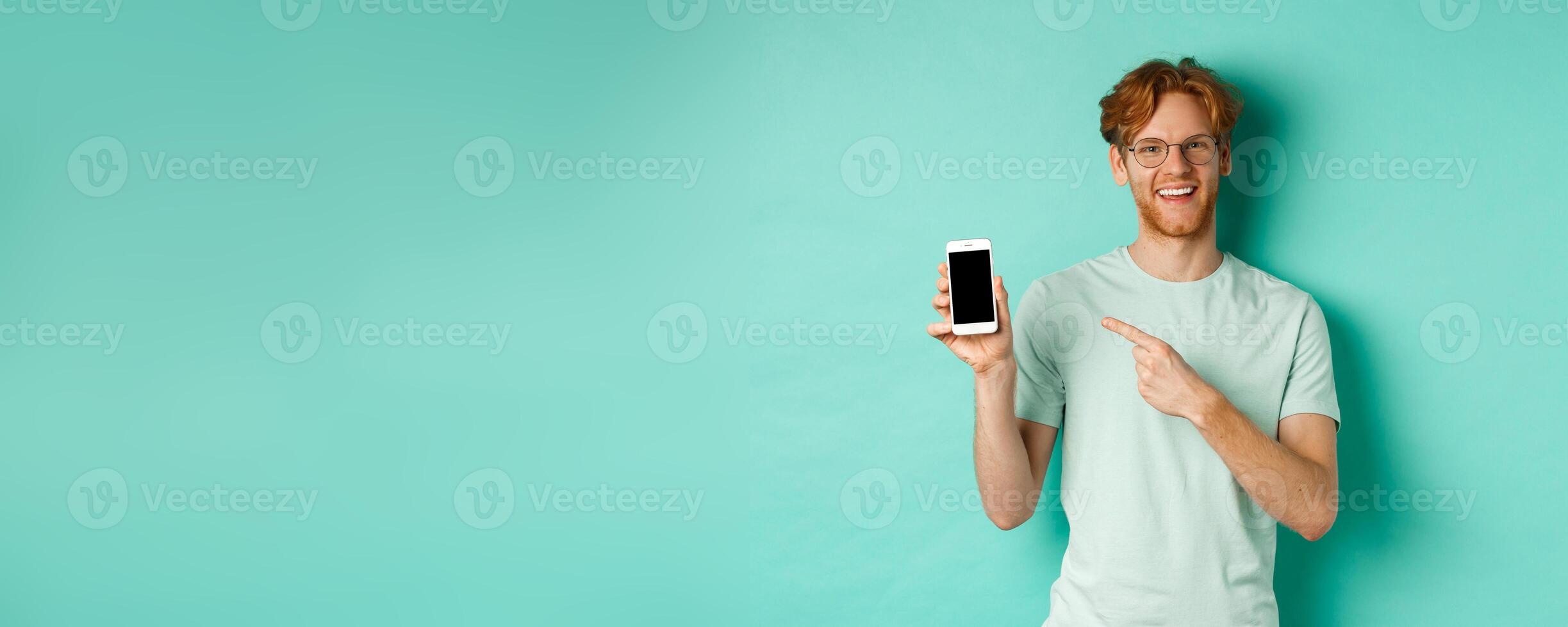 Attractive young man with red beard and hair pointing finger at blank smartphone screen, showing online promotion or app, smiling at camera, turquoise background photo