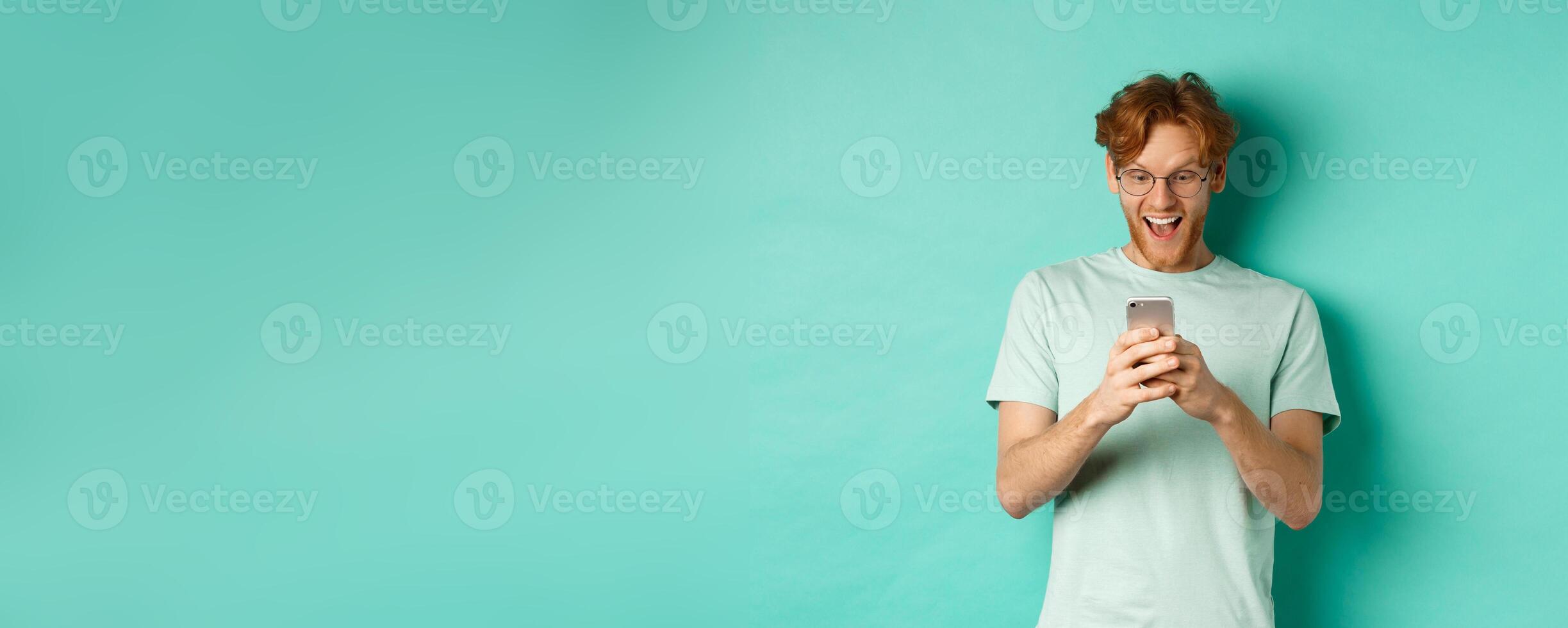 Image of young redhead man in glasses reading phone screen with surprised face, receive amazing promo offer, standing over turquoise background photo