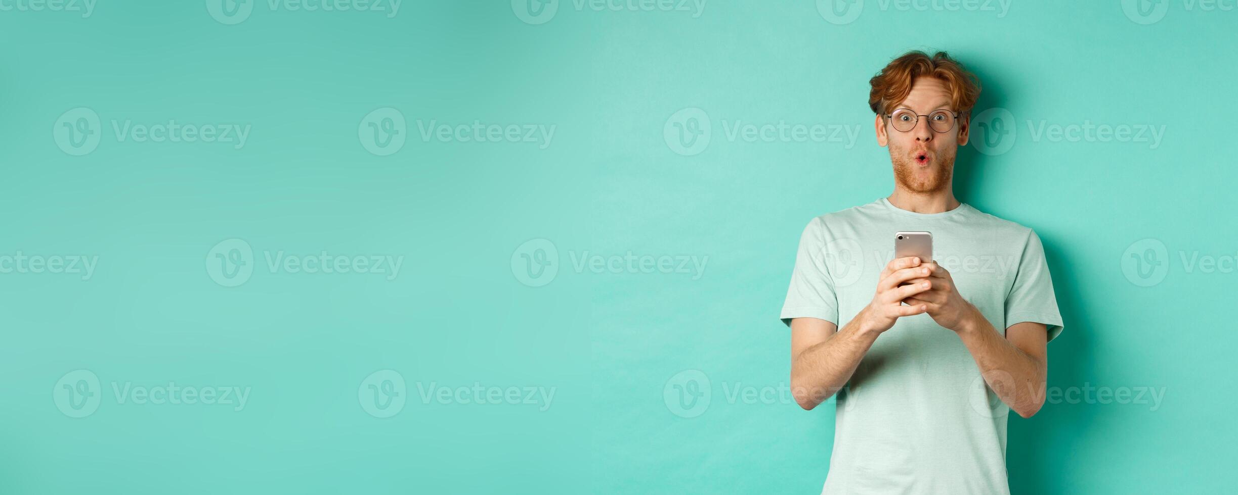 Impressed young man with red hair and glasses, holding mobile phone, saying wow and stare amazed at camera, standing over mint background photo