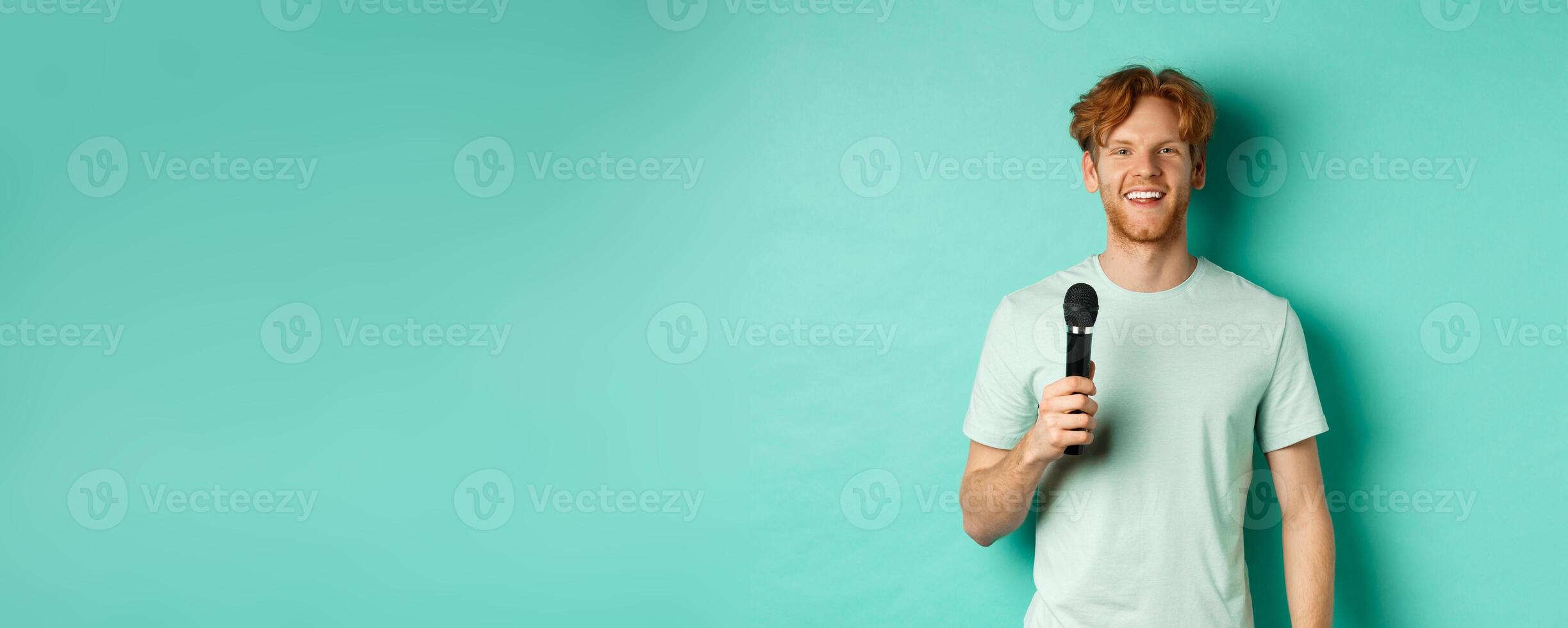 Young redhead man with beard, wearing t-shirt, holding microphone and making speech, singing karaoke, standing over mint background photo