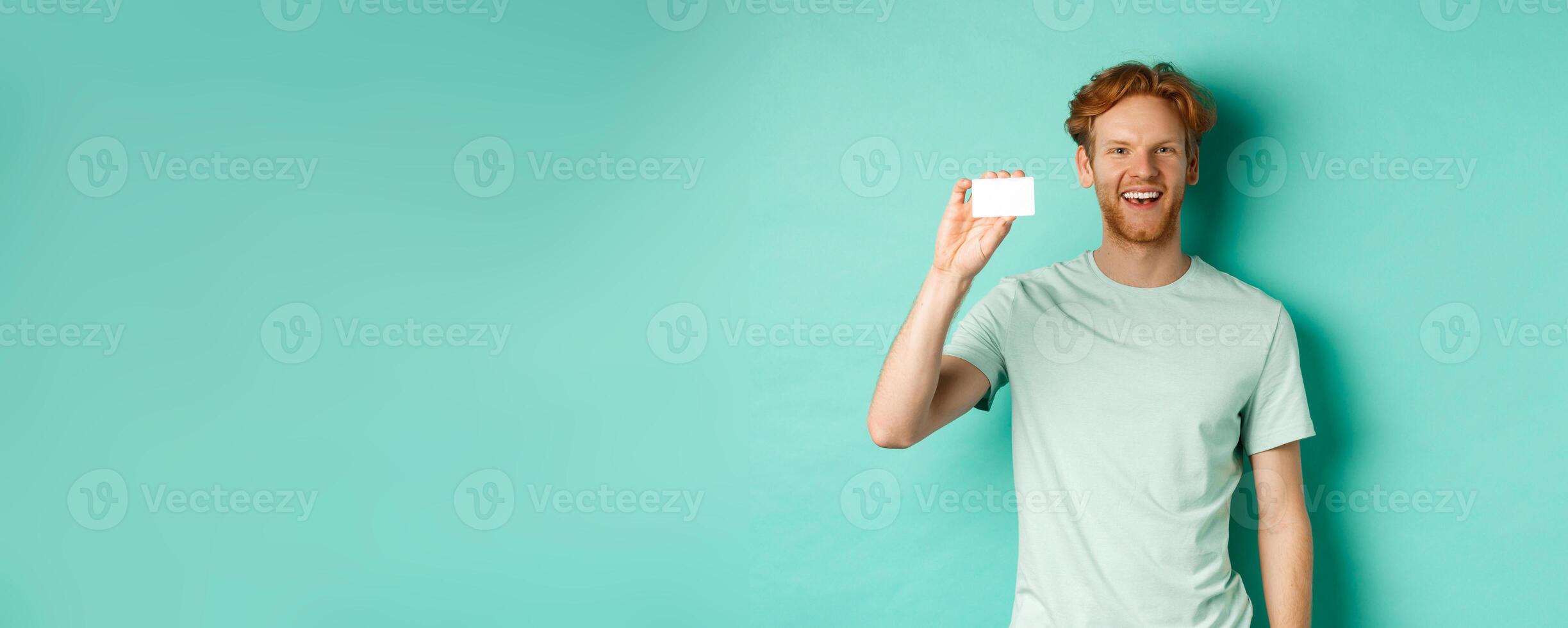 Shopping concept. Cheerful young man in t-shirt showing plastic credit card and smiling, standing over mint background photo