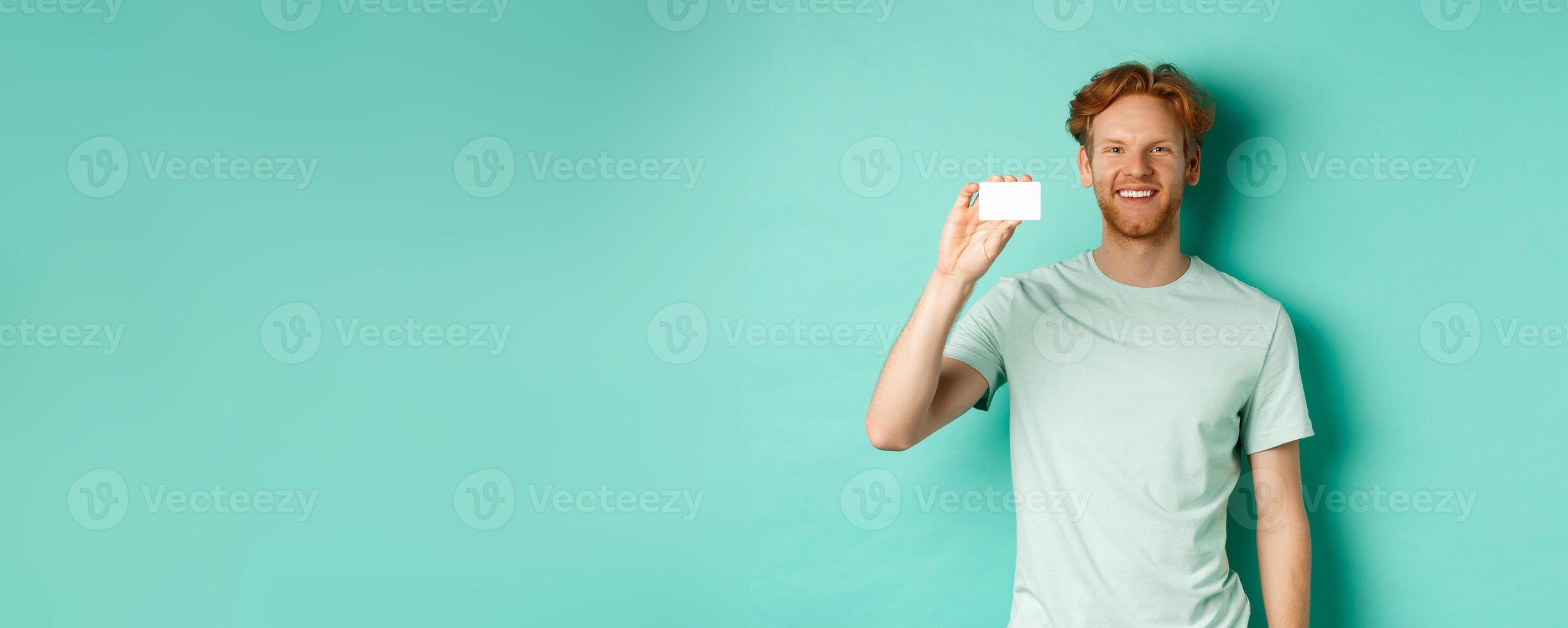 Shopping concept. Handsome redhead man in t-shirt showing plastic credit card and smiling, standing over turquoise background photo