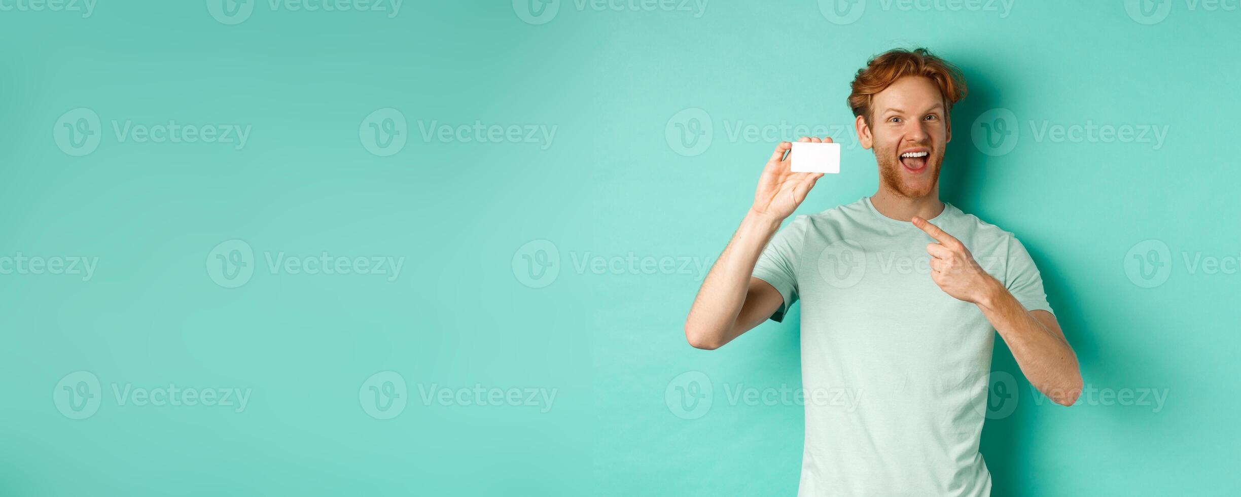 Shopping concept. Handsome redhead man in t-shirt showing plastic credit card and smiling, standing over turquoise background photo