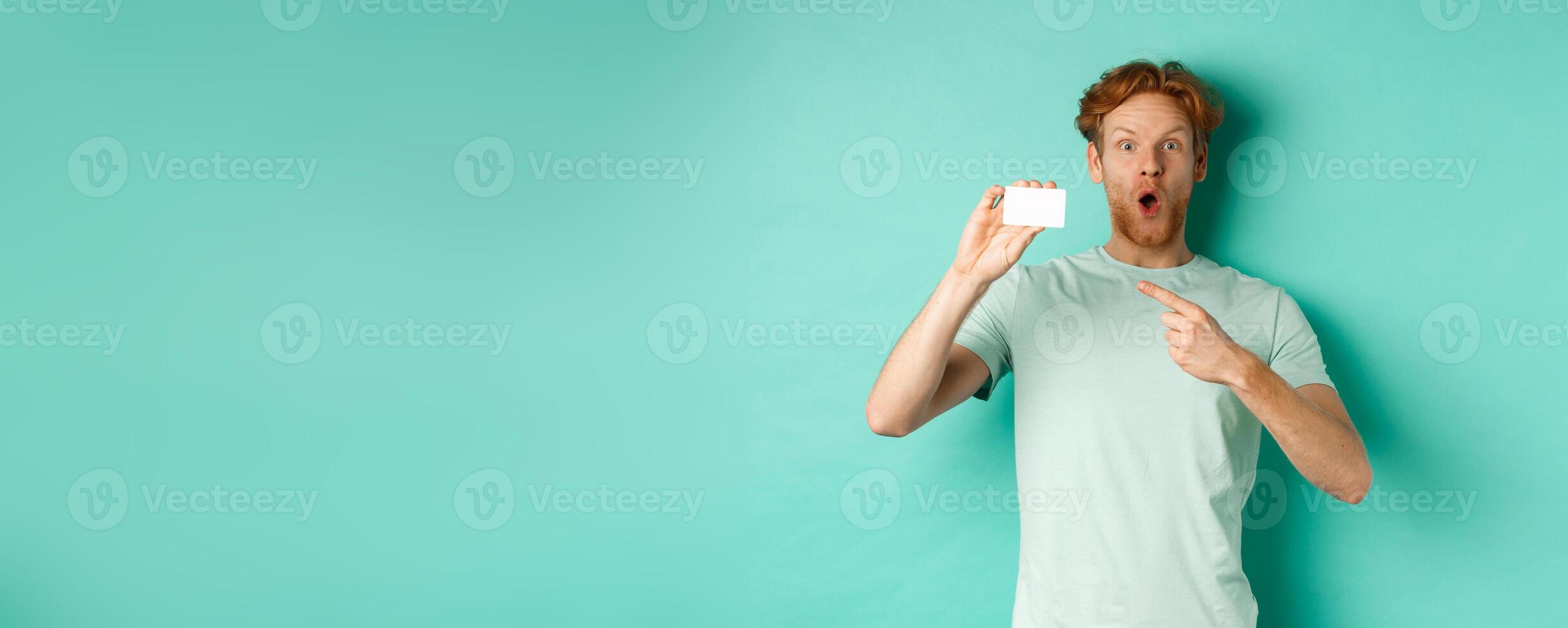 Shopping concept. Handsome redhead man in t-shirt showing plastic credit card and smiling, standing over turquoise background photo