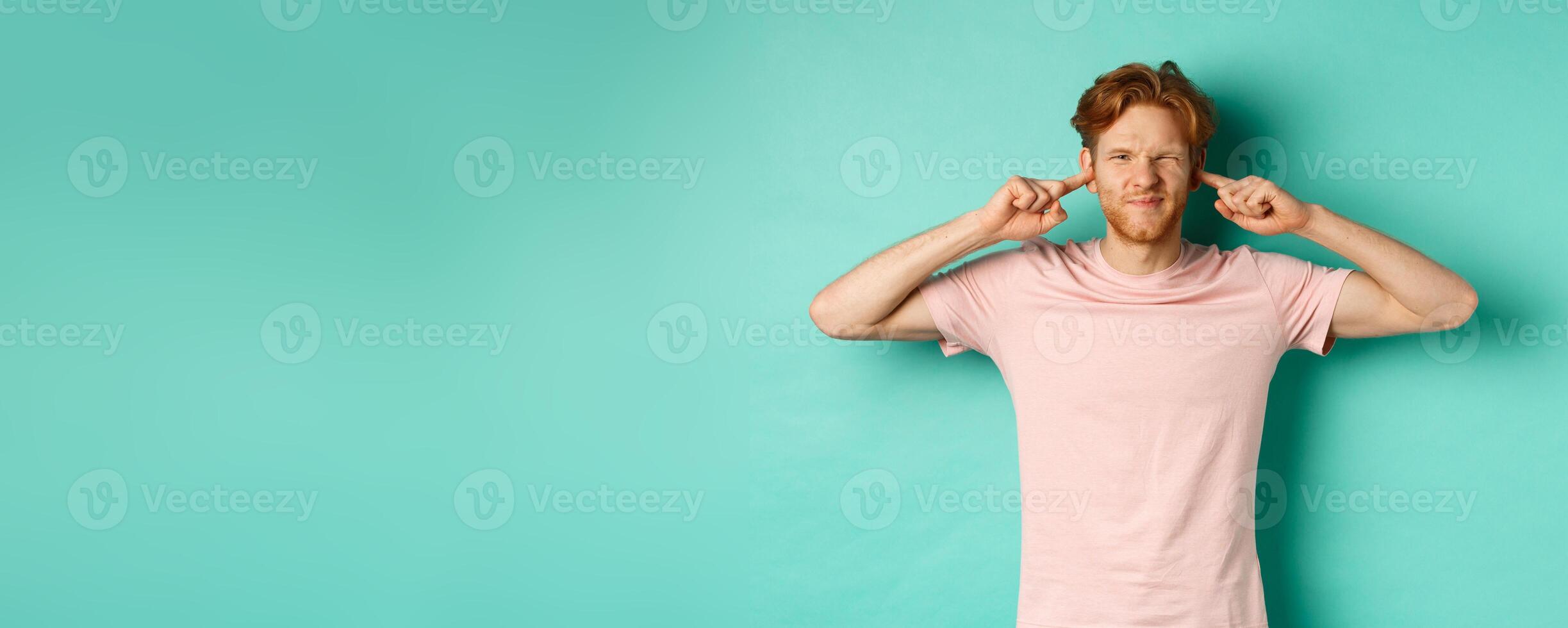 Annoyed young man with red hair and beard shut his ears and grimacing, disturbed by loud bothering sound, noisy neighbours, standing over turquoise background photo