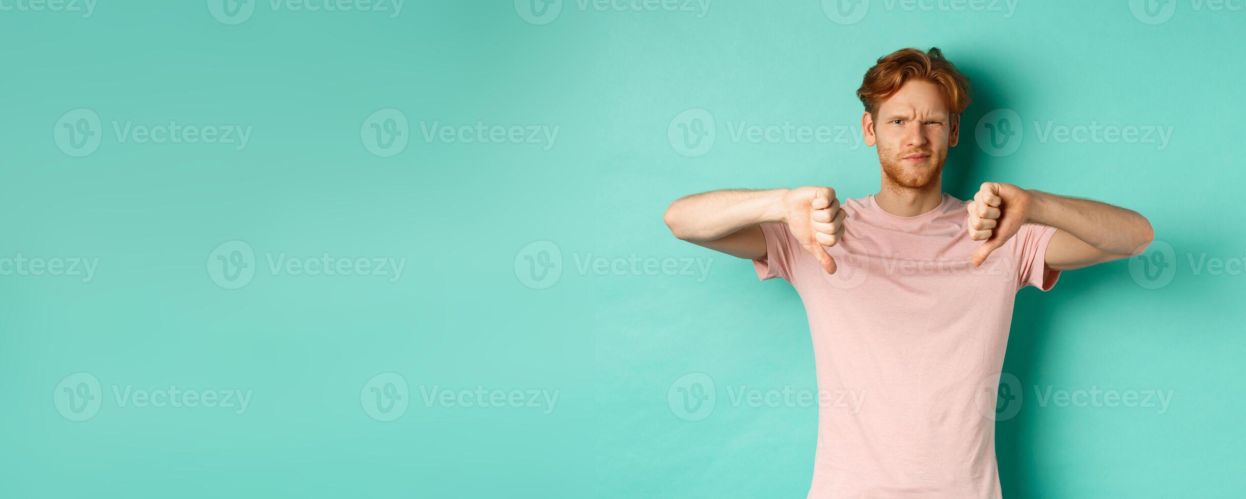 Disappointed guy with red hair showing thumbs-down, frowning and looking skeptical, epxress dislike and disapproval, standing over turquoise background photo